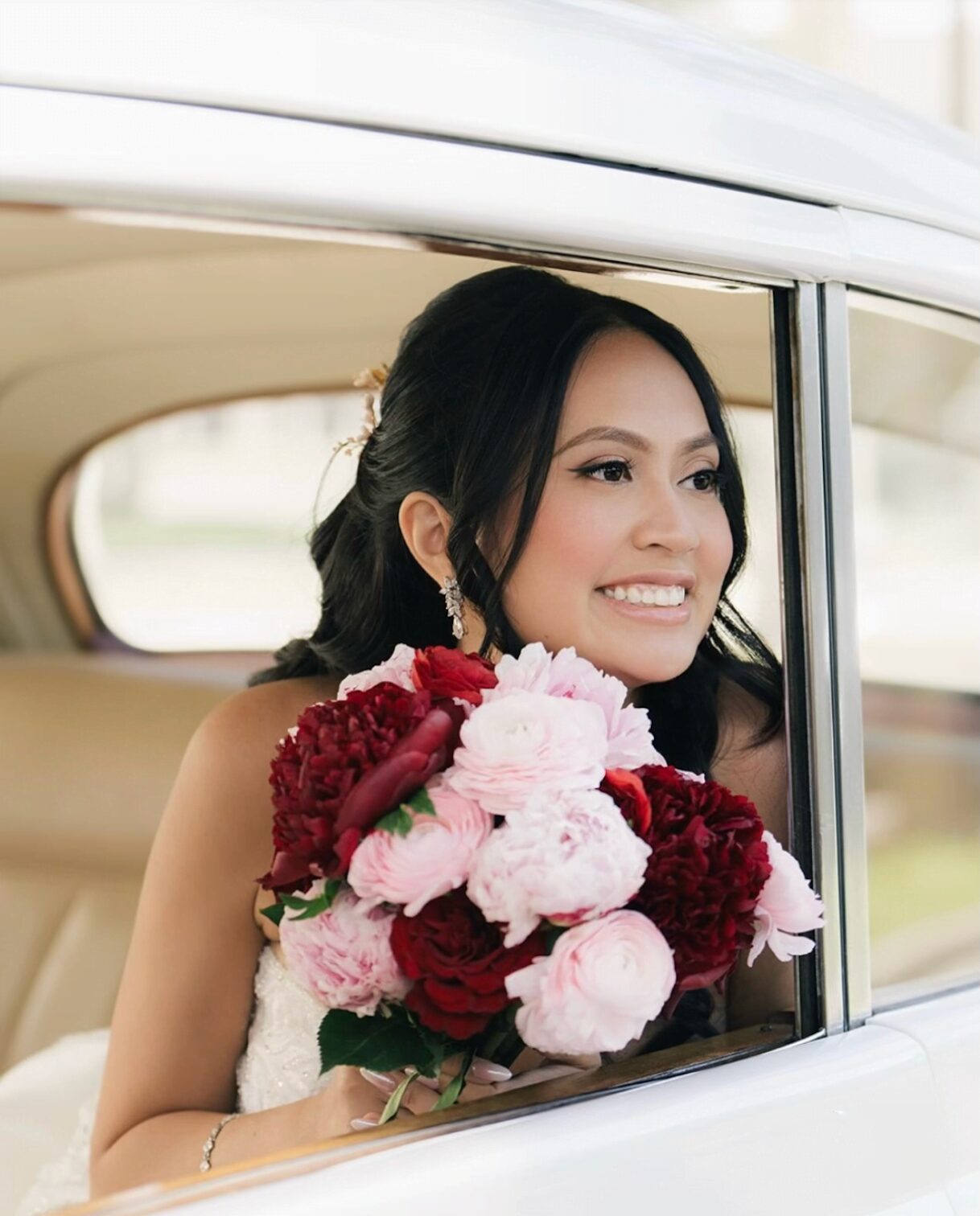 Young woman with dark hair smiling, holding a bouquet of pink and red flowers, sitting in a vehicle window.