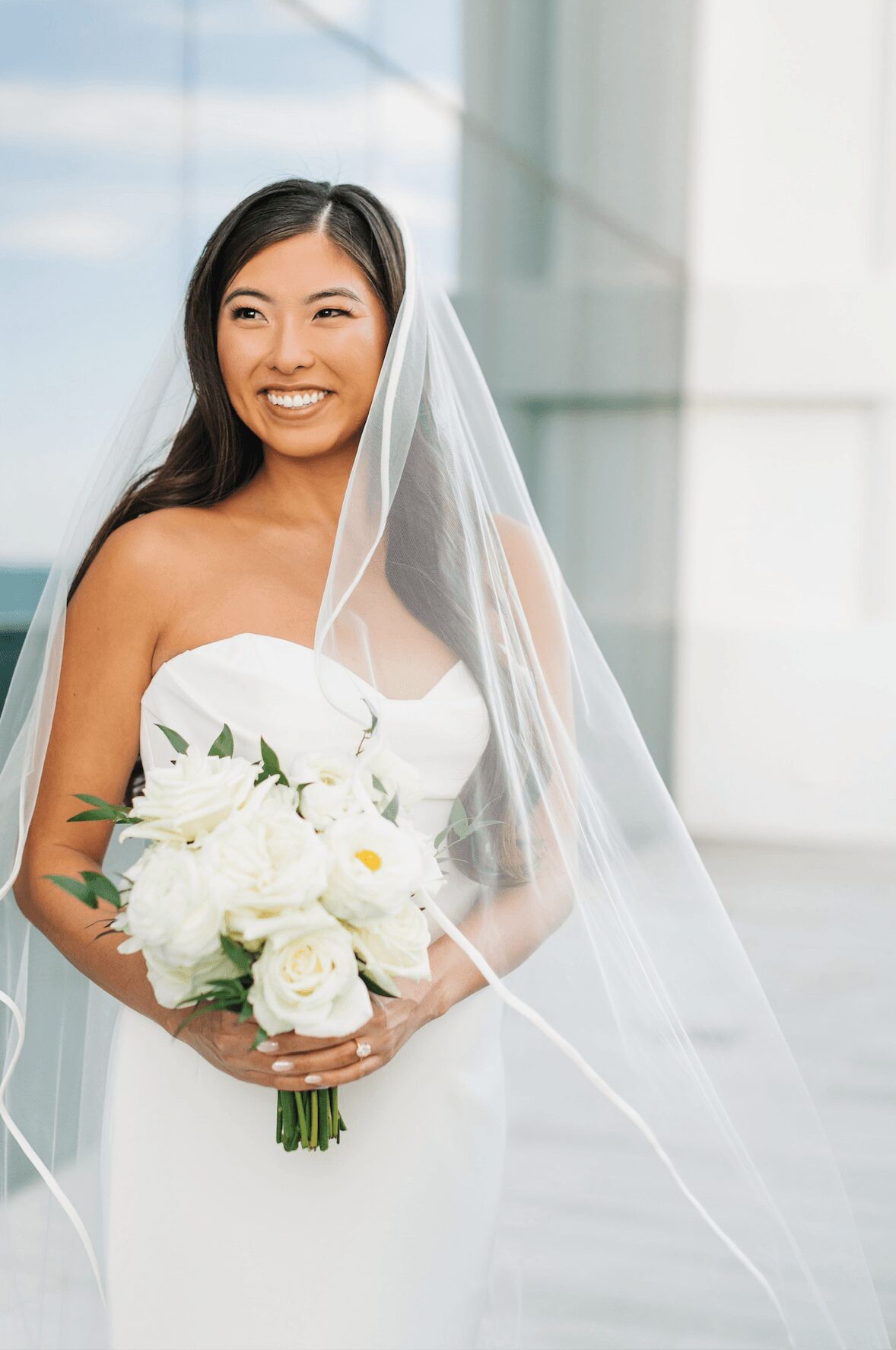 Smiling woman in wedding dress holding a bouquet of white flowers, with a veil and modern building background.