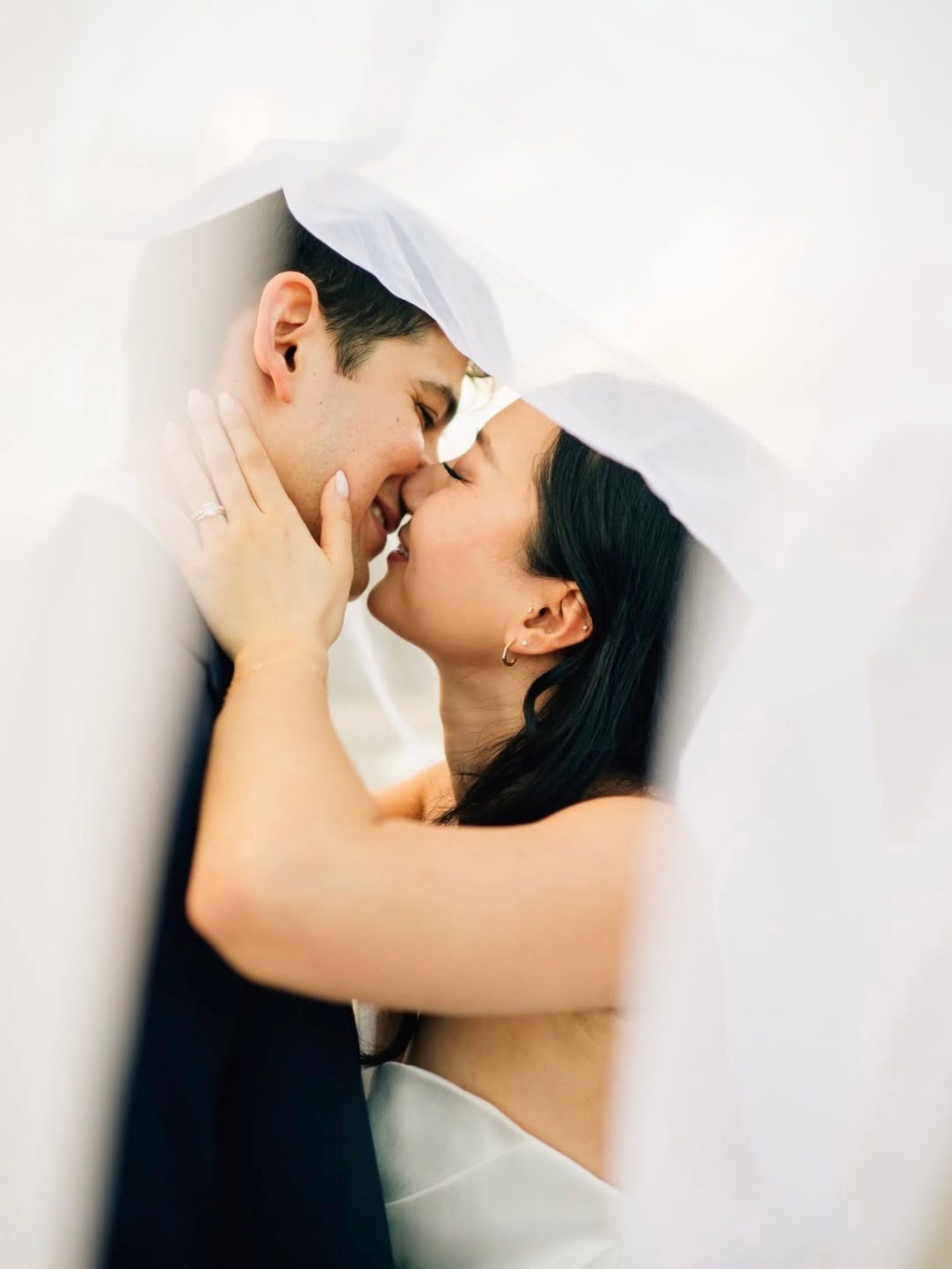 Couple with faces close, smiling, touching noses, under white fabric, intimate moment, woman with dark hair, man with short dark hair