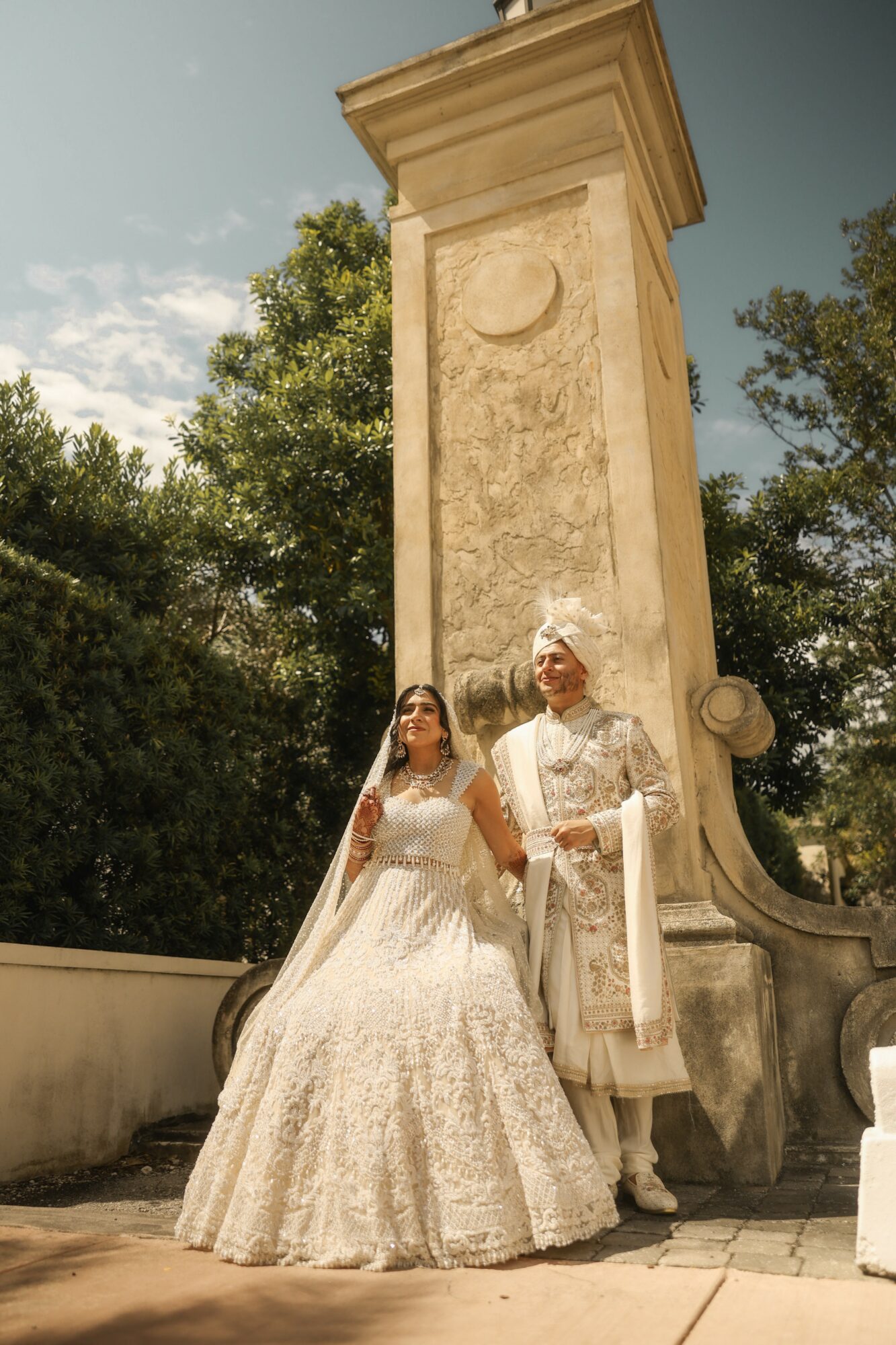 Bride in wedding dress and groom in traditional attire stand outdoors near a tall stone monument with trees in background.
