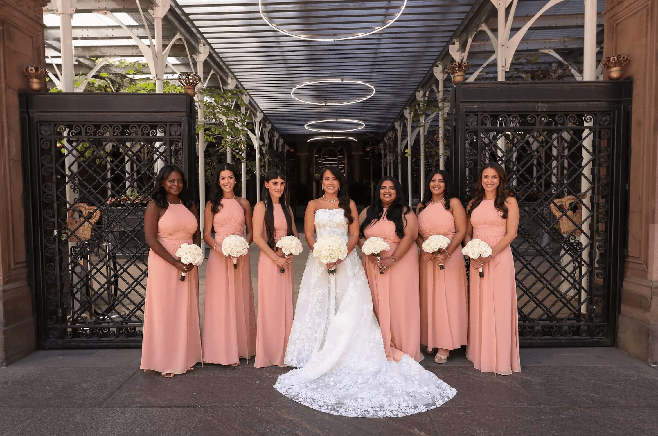 Seven women in pink dresses and a bride in a white gown stand in front of a decorative gate, holding bouquets.
