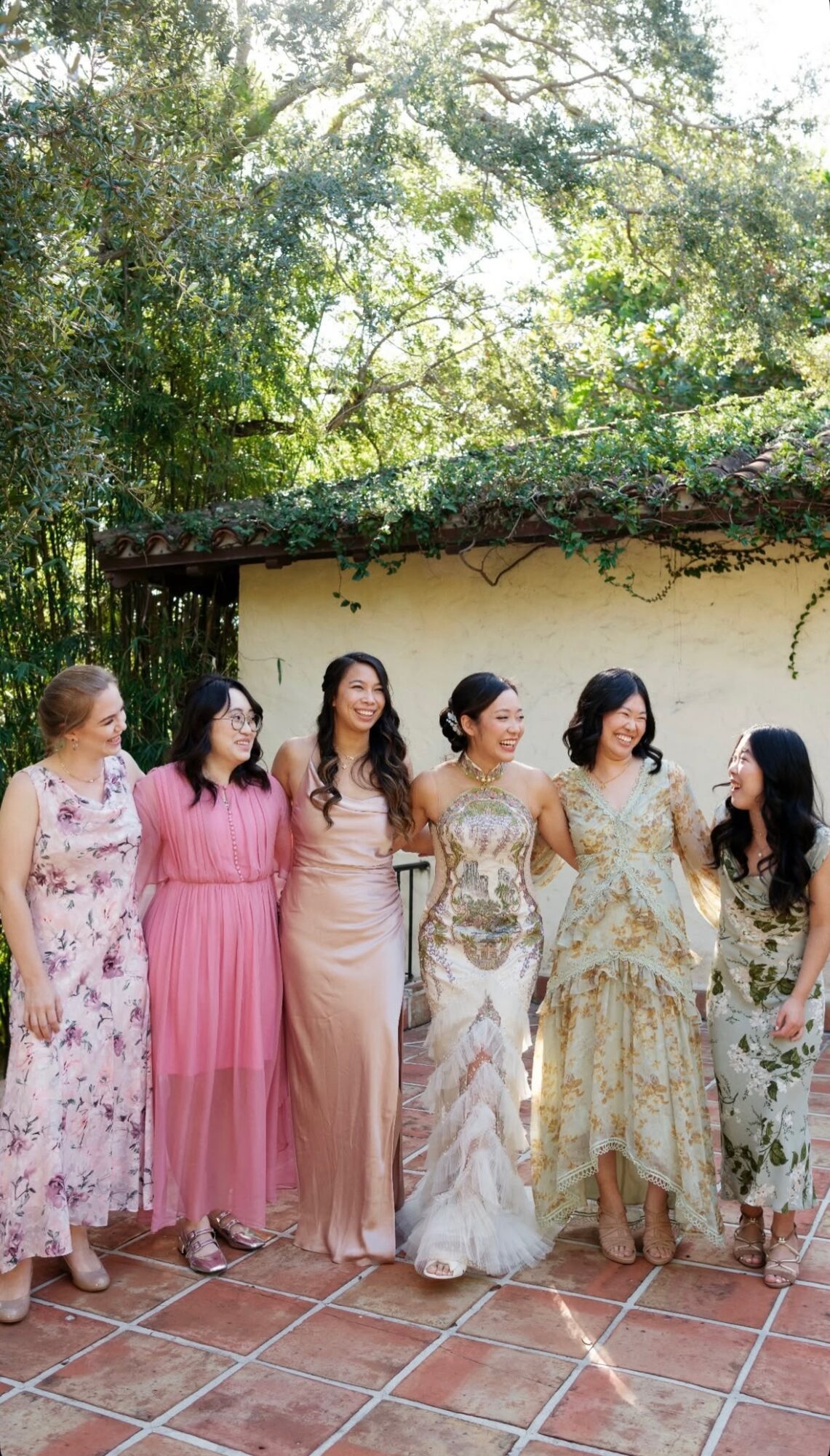 Six women in colorful dresses standing outdoors on a tiled surface, with trees and a wall in the background.