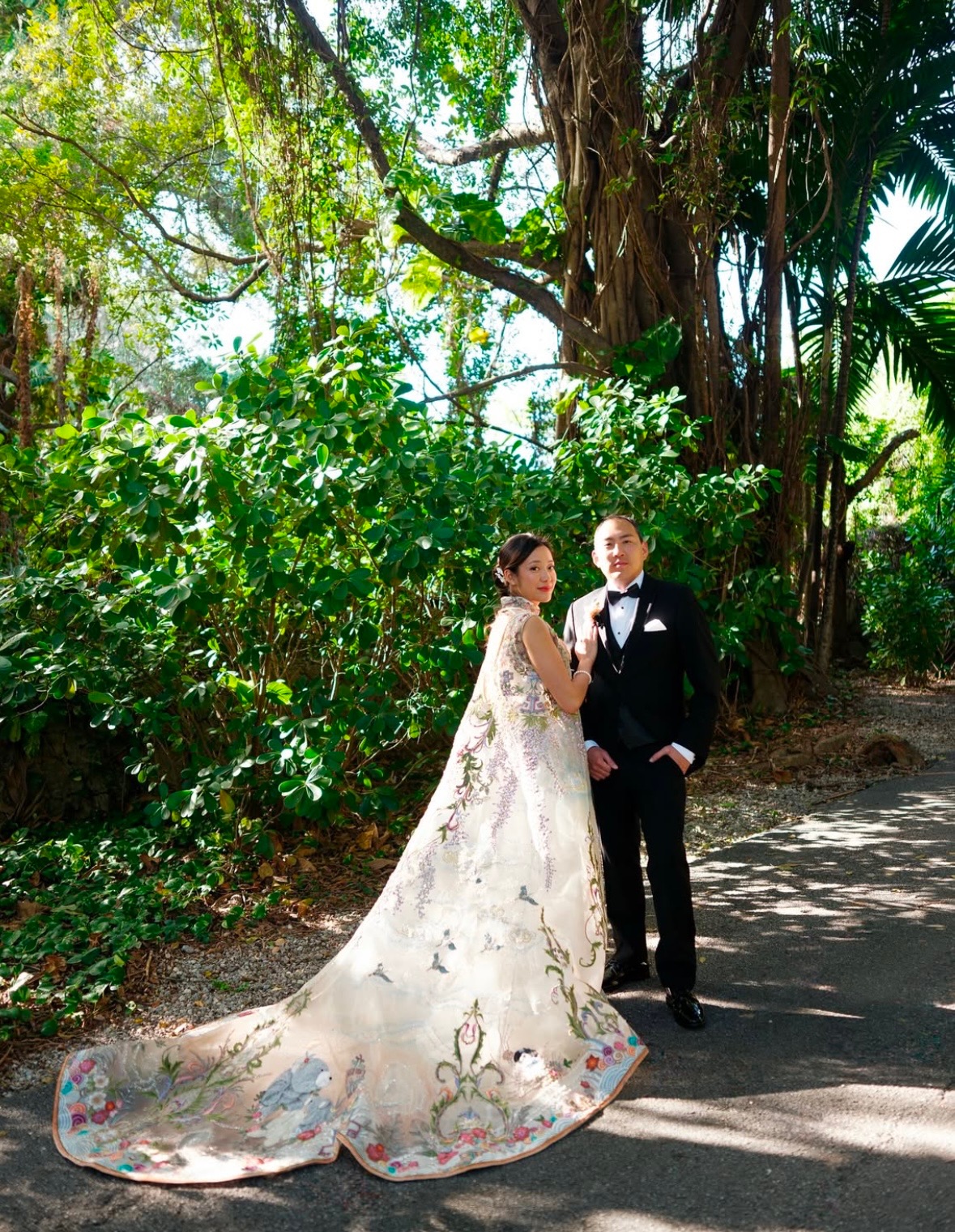 A bride and groom stand on a path surrounded by lush green trees and plants, sunlight filtering through leaves.