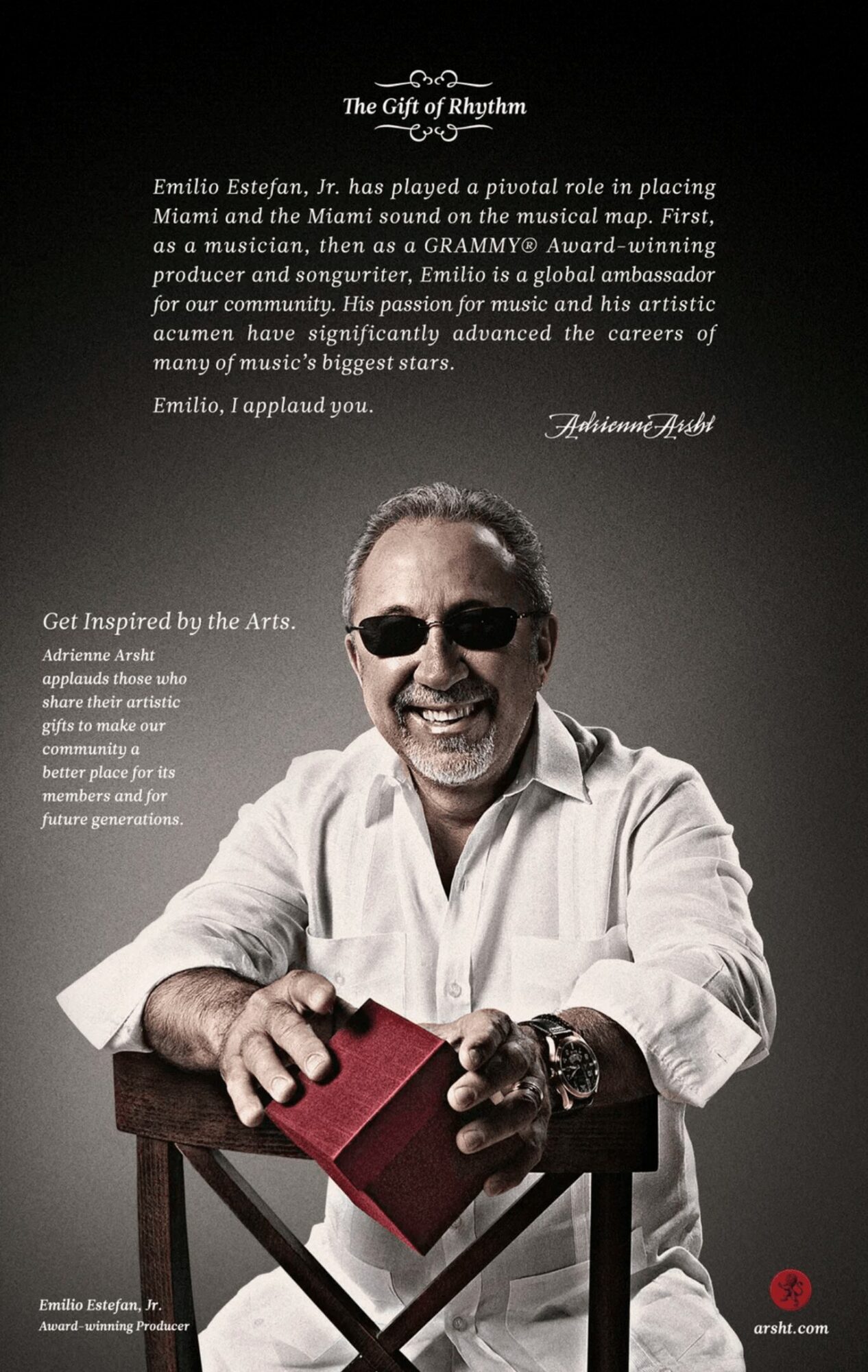 Smiling man wearing sunglasses and a white shirt, holding a red box, sitting at a wooden table.