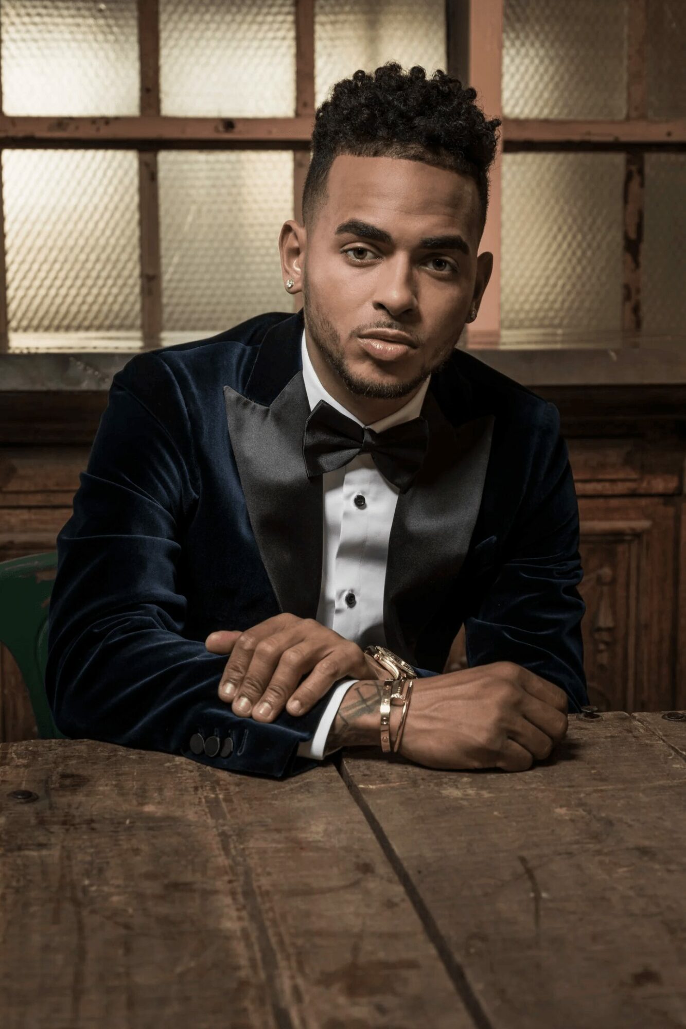 Young man in tuxedo with bow tie sitting at a wooden table, background of window panes, serious expression.