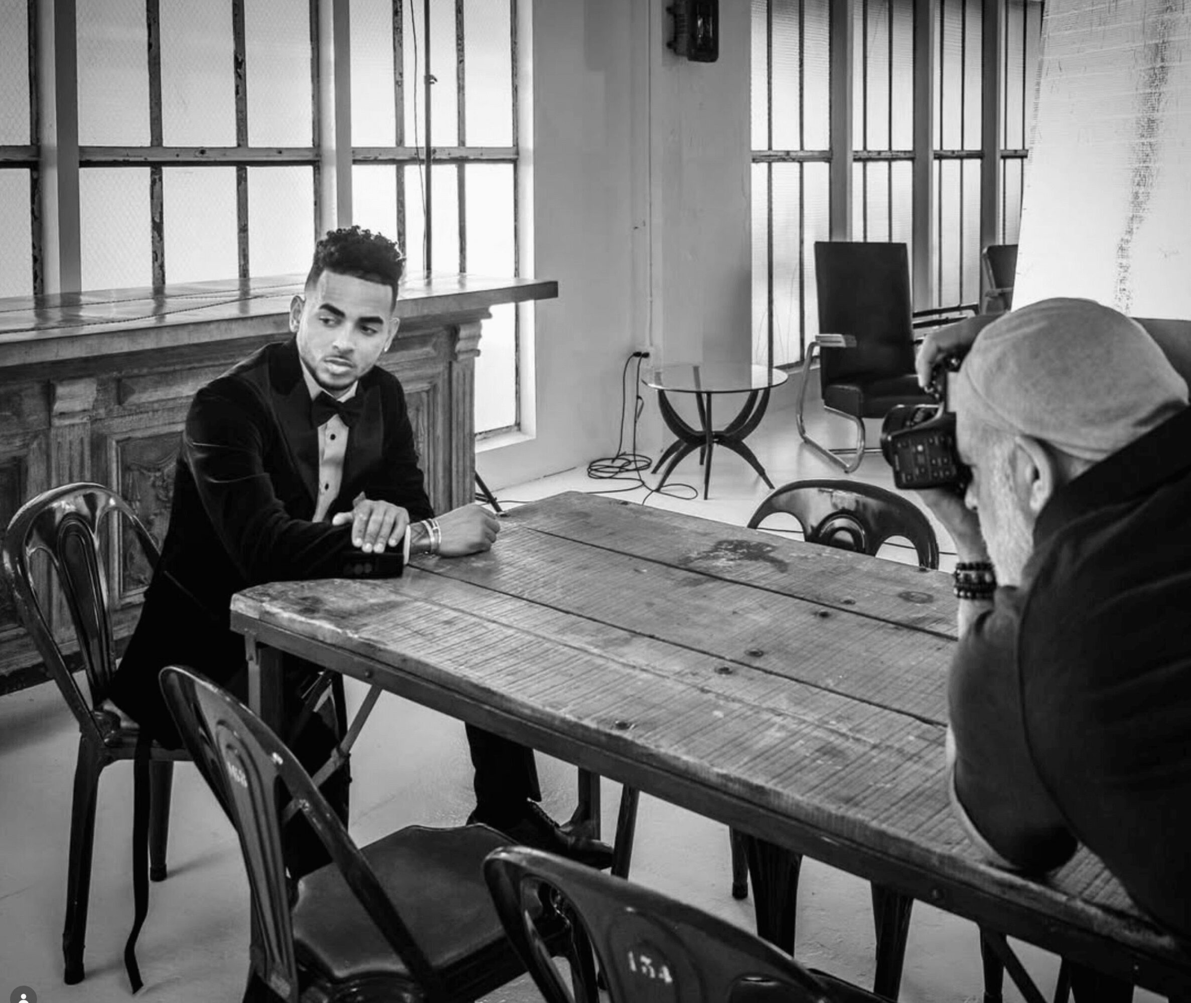 Young man in a suit sitting at a wooden table, facing an older man with glasses in a room with large windows.