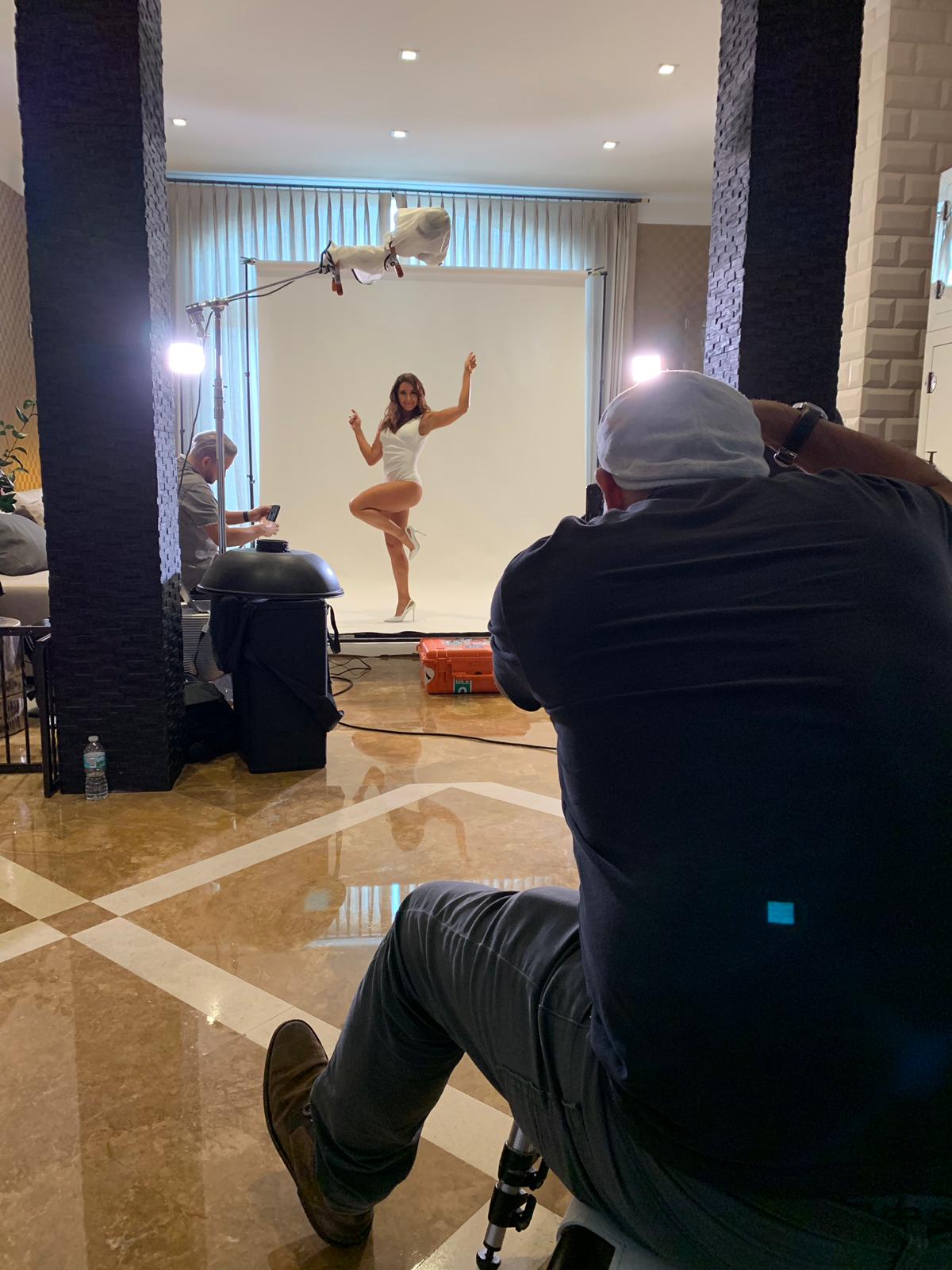 Photographer taking picture of a dancer in a studio with a white backdrop, two bright lights, and a person in the foreground wearing a cap.