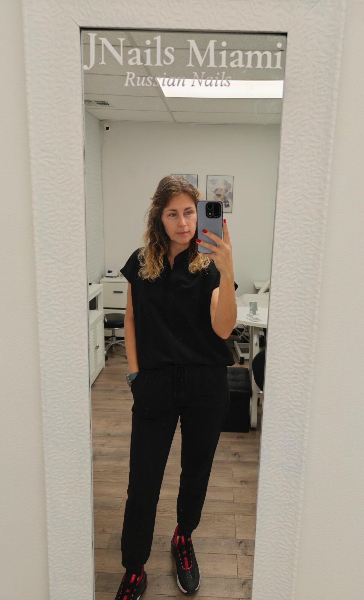 Young woman with wavy hair taking a mirror selfie in a nail salon, wearing a black outfit and sneakers.