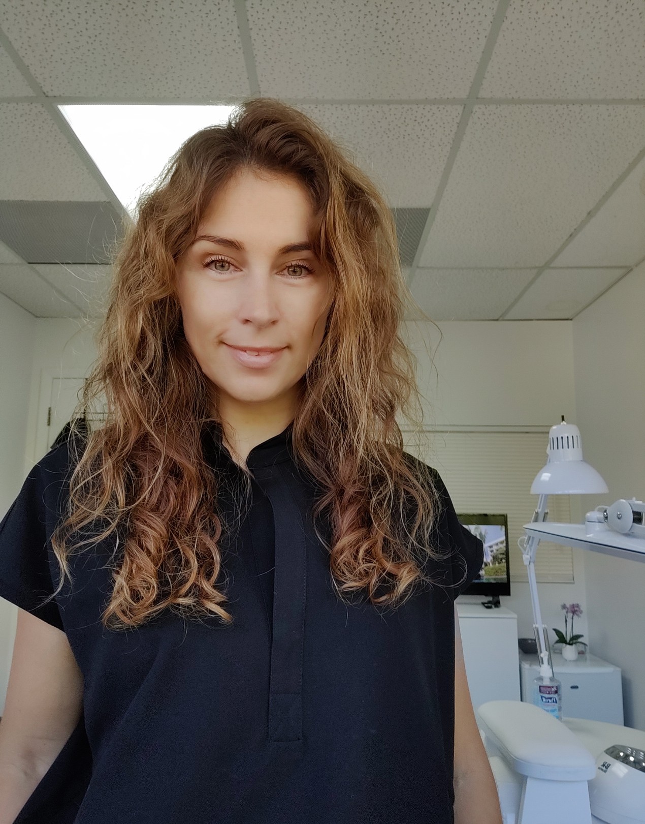 Young woman with curly hair smiling in an indoor setting, wearing a dark shirt, with ceiling lights and office equipment in background.