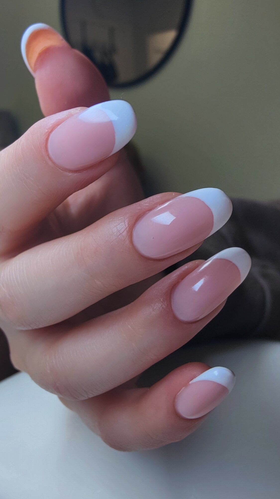 Hand with nails painted in French manicure style, featuring white tips and natural pink base, held against a blurred background.