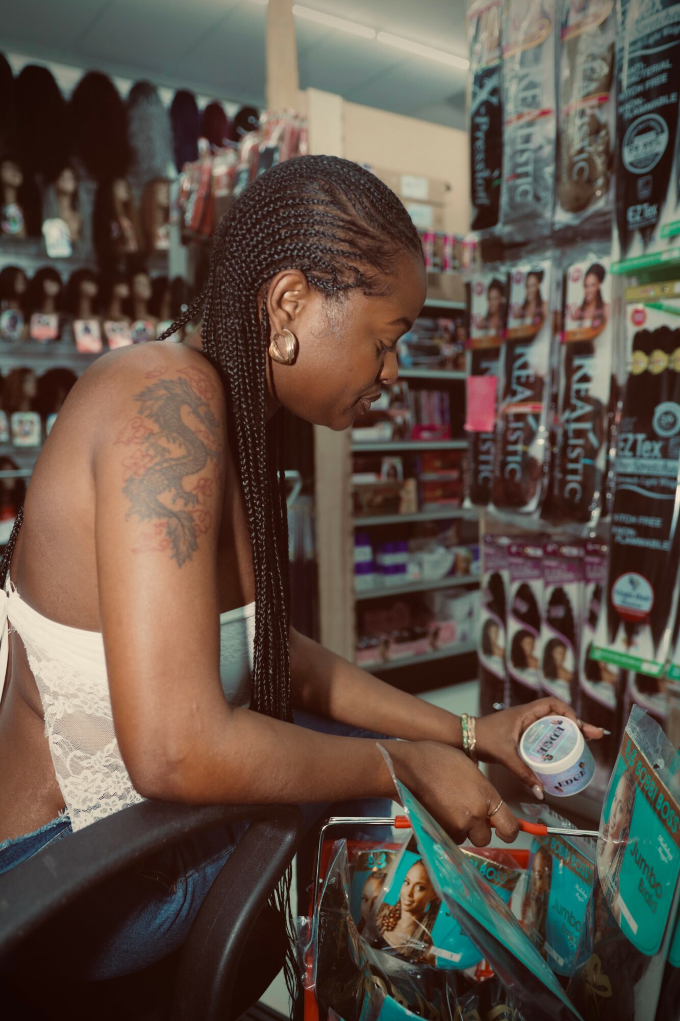 Woman shopping in store, looking at hair products on shelf, with braided hair and tattoos on shoulder, shopping cart nearby.