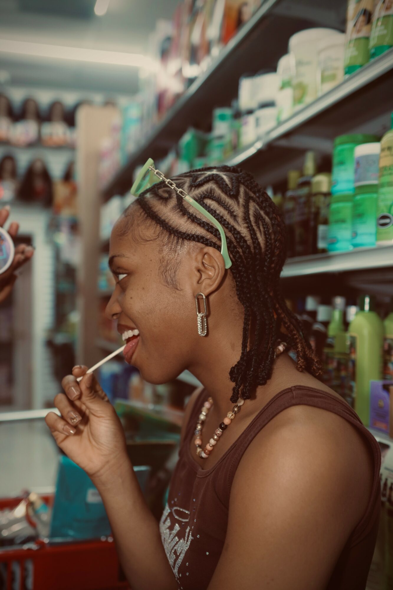 Woman with braided hair and earrings in store aisle, smiling and holding a lollipop, shelves with products behind her.