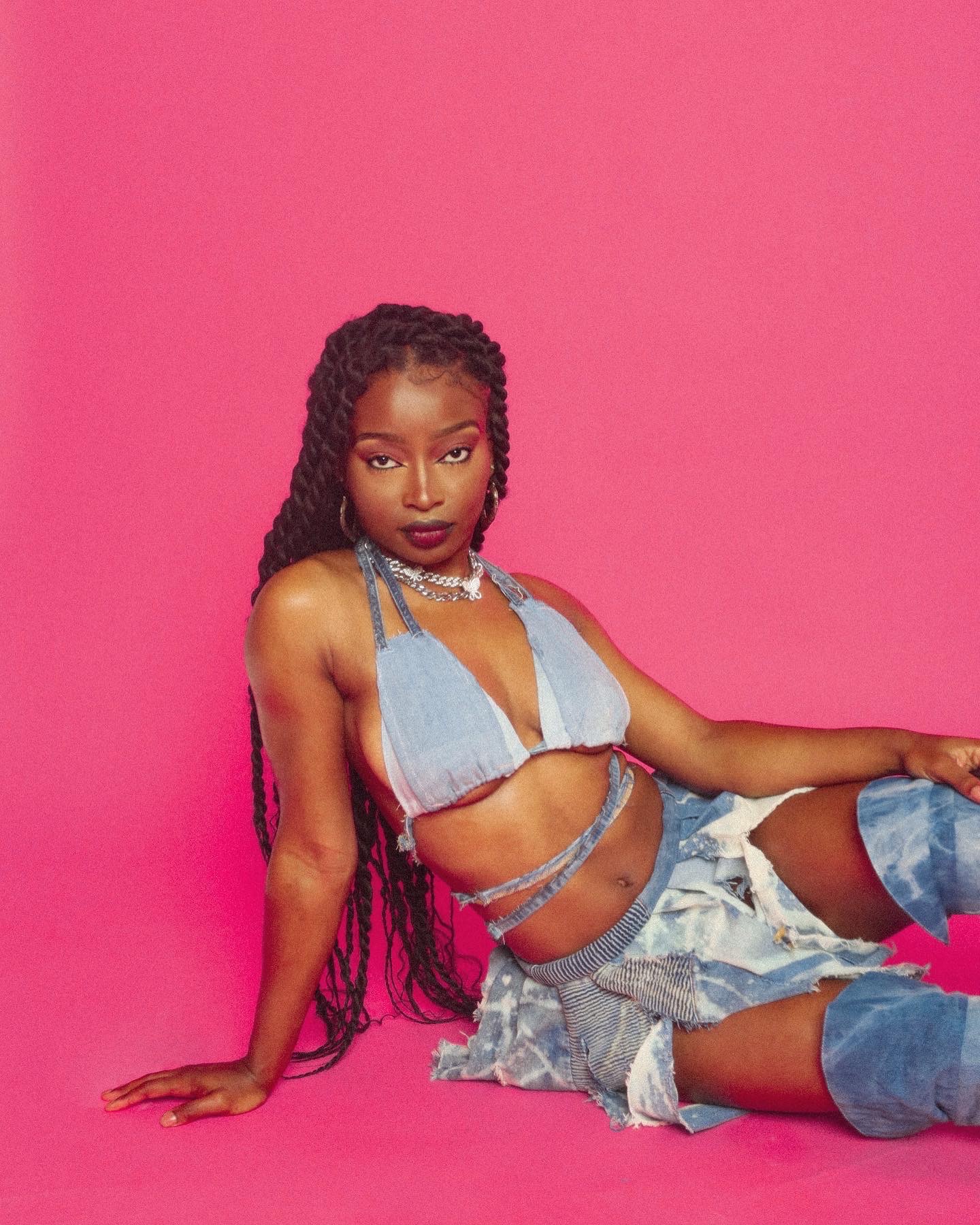 Young woman with long braids poses against pink background, wearing denim outfit and jewelry.