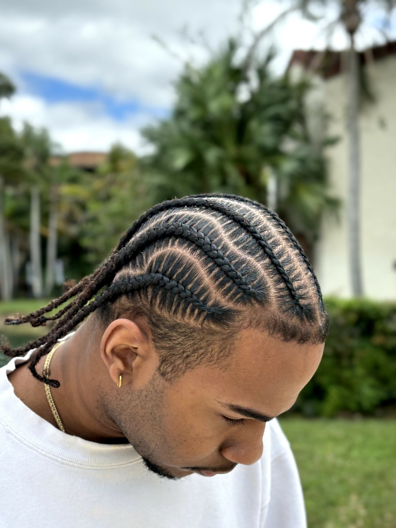 Young man with braided hairstyle looking downward outdoors, greenery and a building in background.