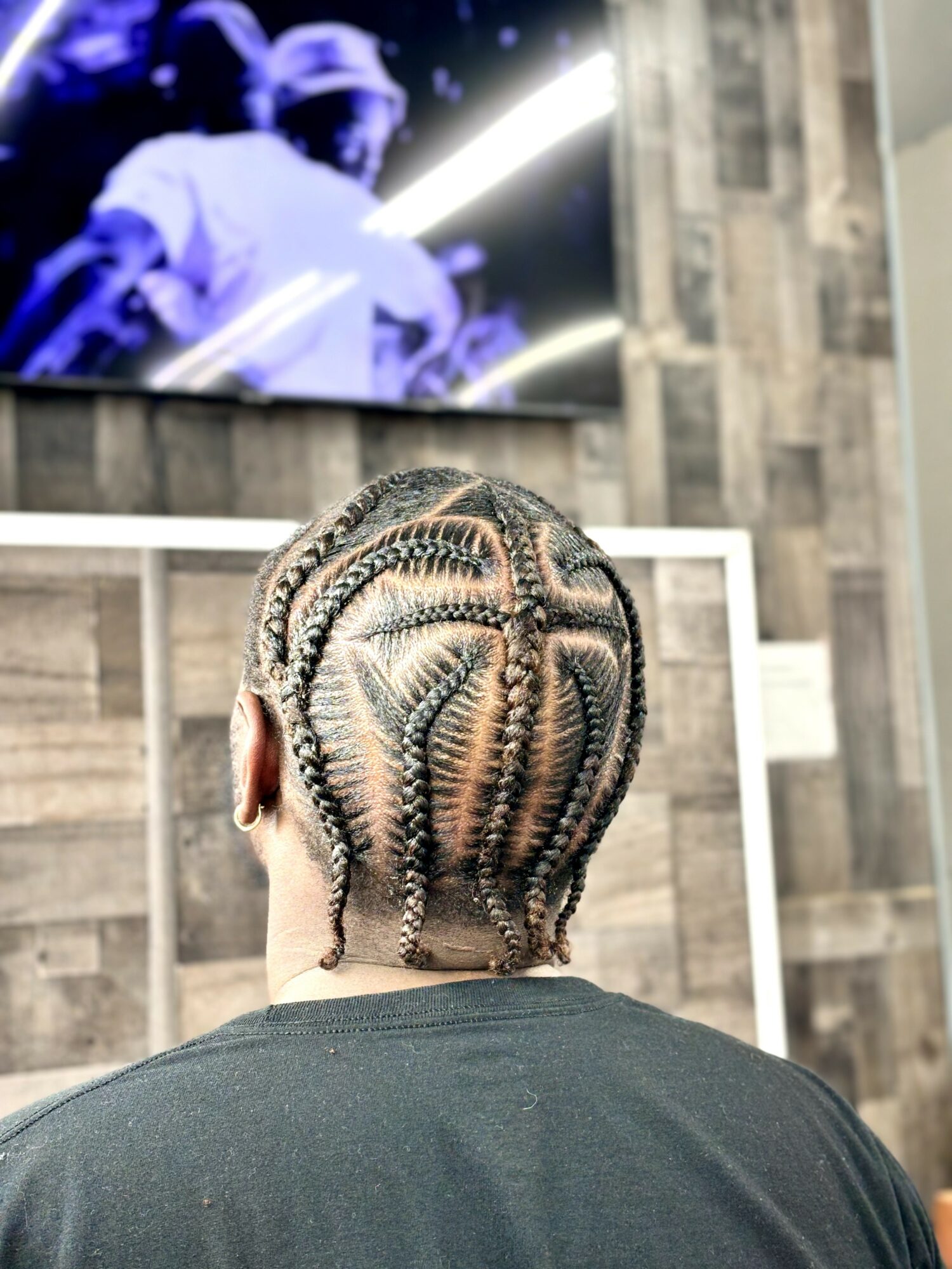 Back of a person's head with braided hair, in a room with a large screen showing a person in a white outfit, wall with wood paneling