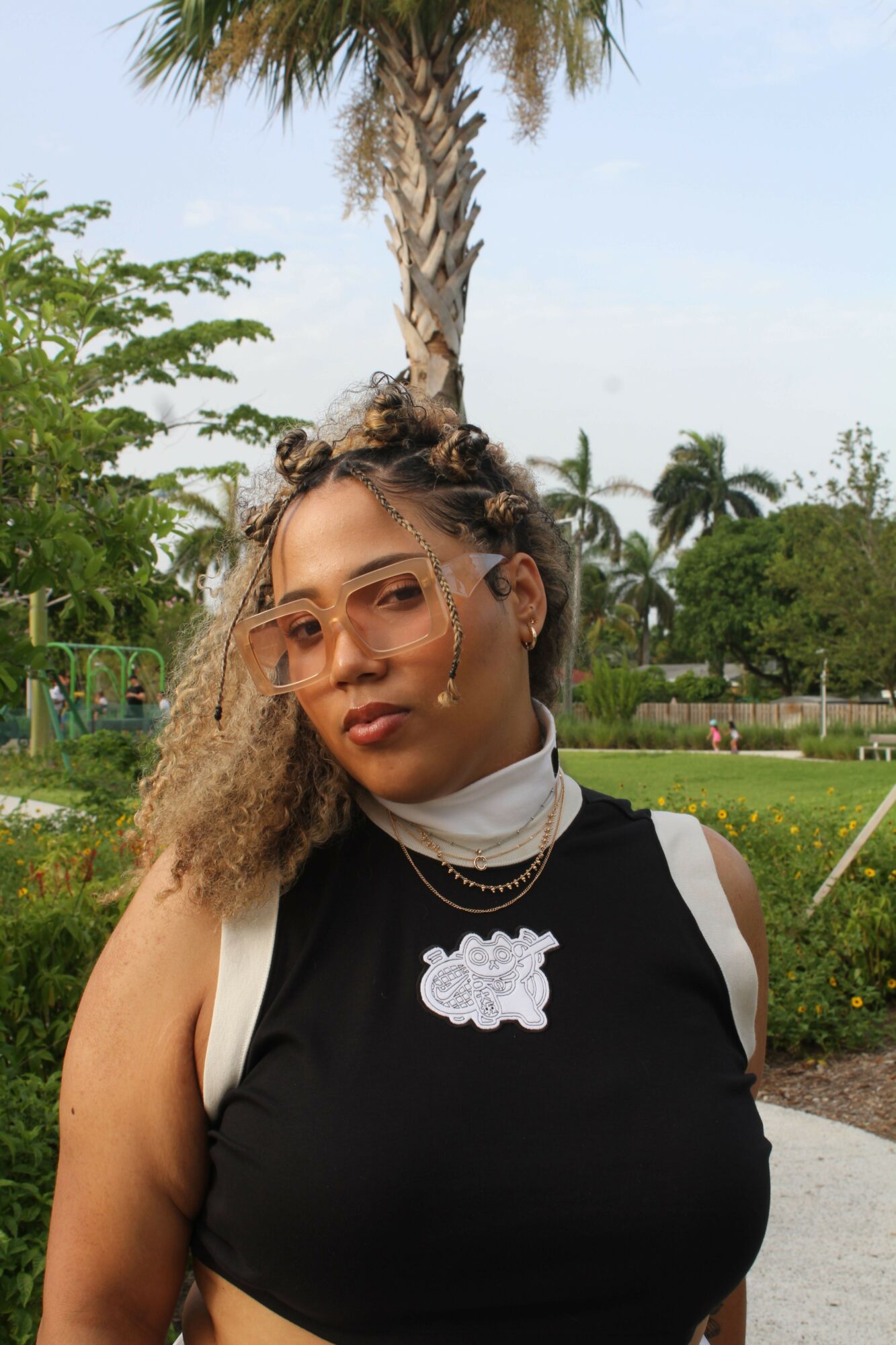 Young woman with curly hair and glasses outdoors, wearing a black and white top, standing in a park with trees and sky.