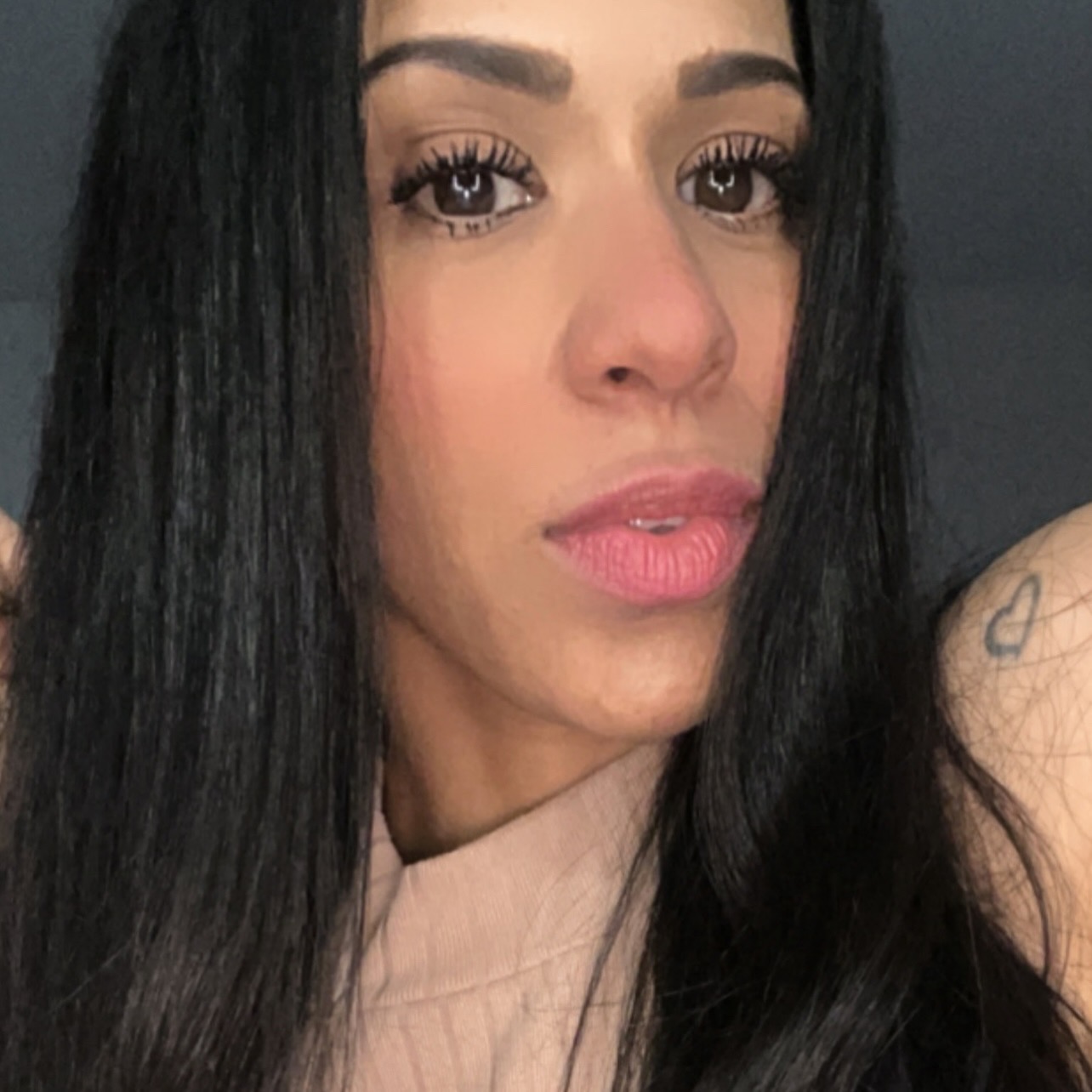 Close-up of a woman with long dark hair, makeup, and a tattoo on her shoulder, looking at the camera.