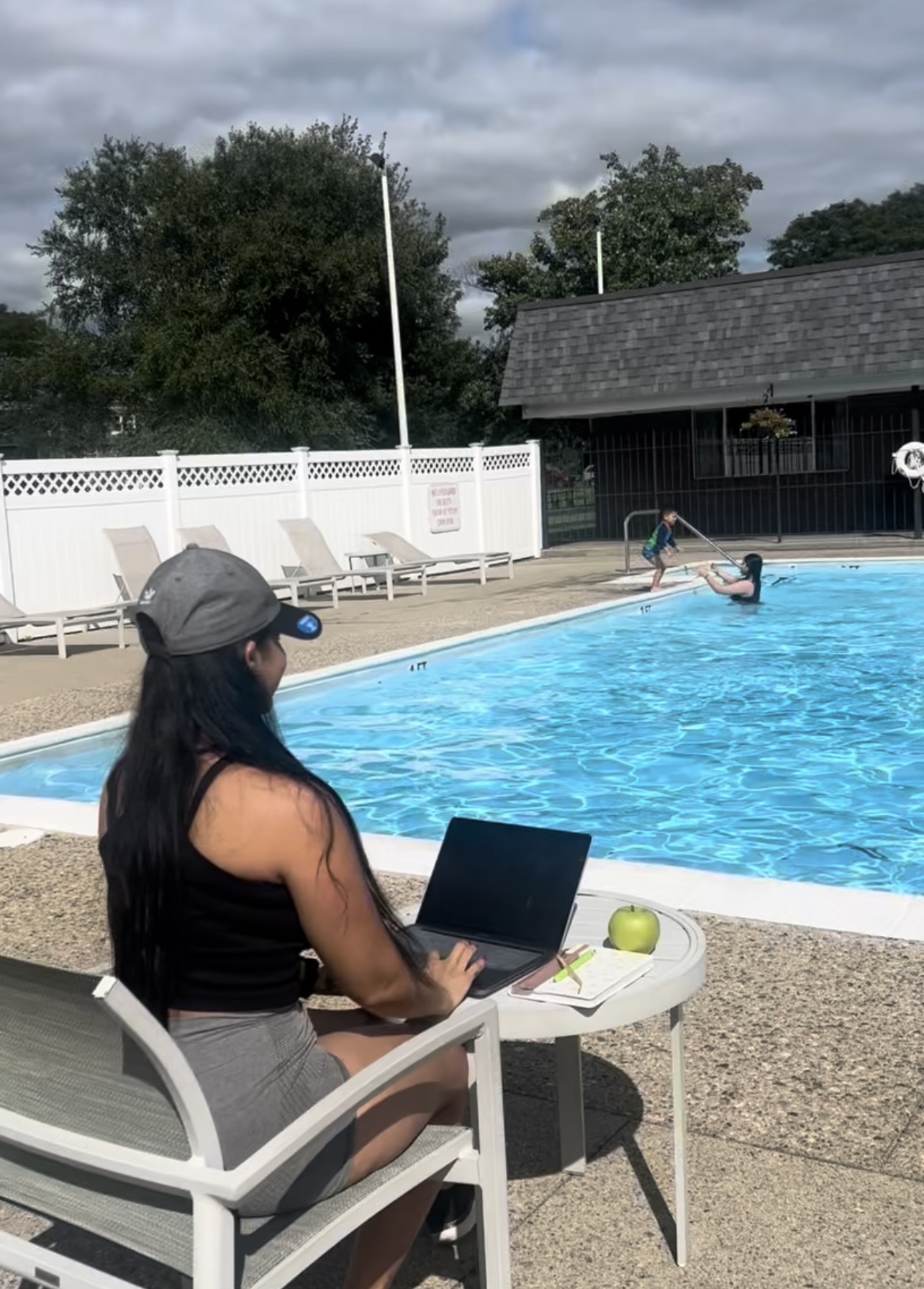 Woman sitting at a table with a laptop near a swimming pool, with a person swimming in the pool, trees, and a building in the background.
