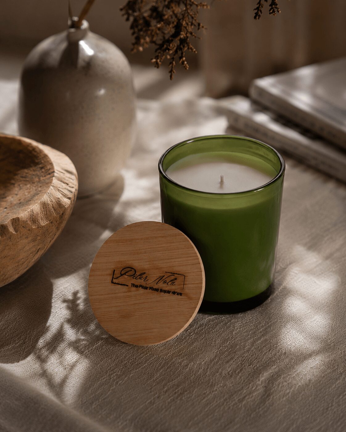 Green candle in a glass holder, wooden lid with text, ceramic vase, wooden bowl, and books on a table.
