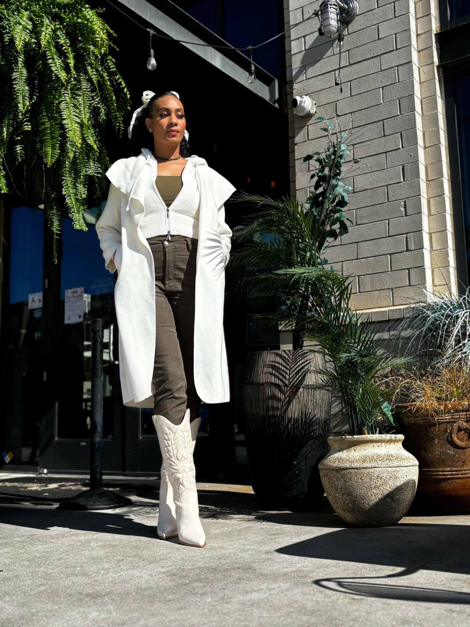 Woman standing outdoors in sunlight, wearing a white coat, black pants, and white boots, with potted plants nearby.