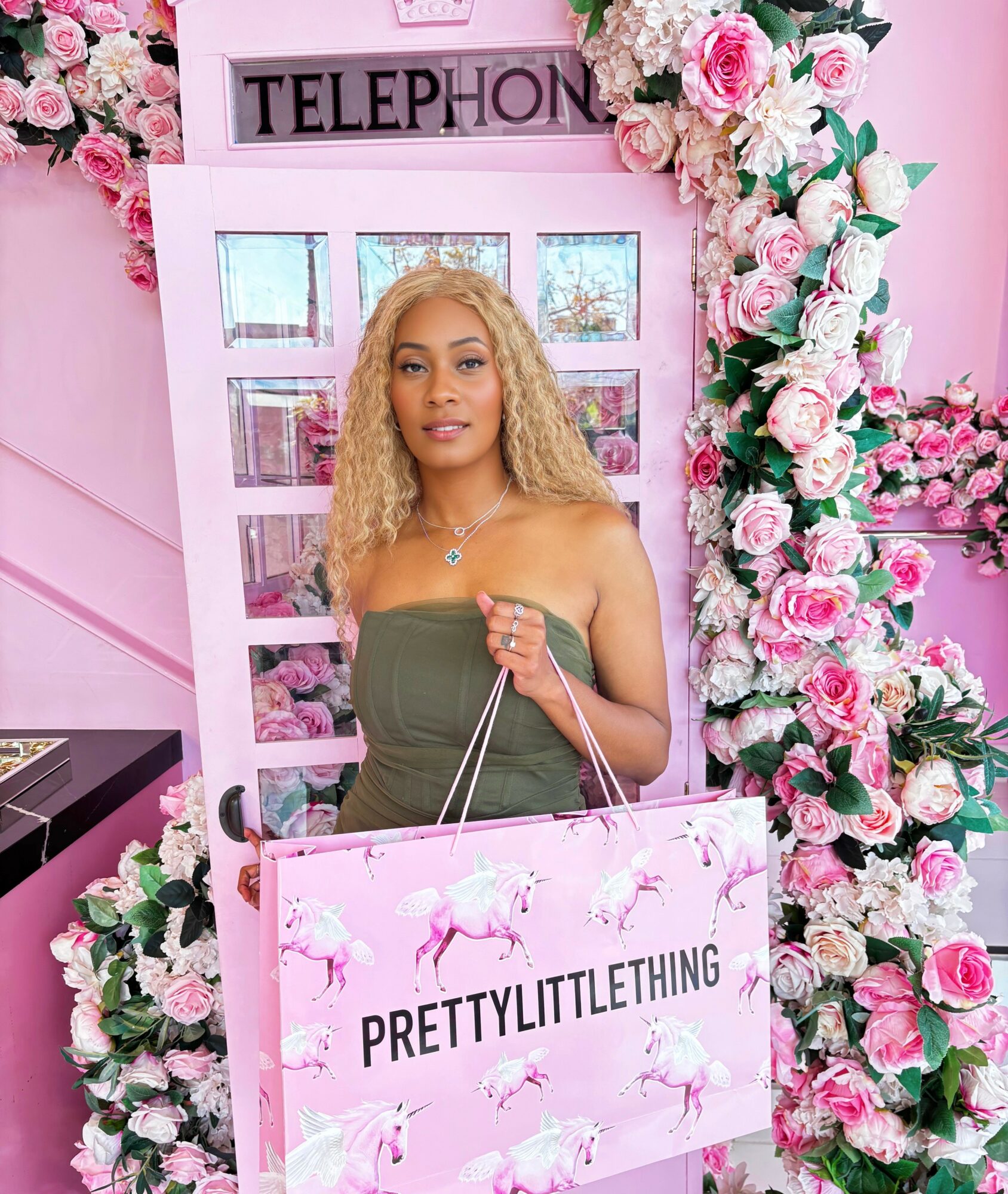 Woman with curly blonde hair in green dress holding a pink shopping bag in front of a pink phone booth decorated with flowers.