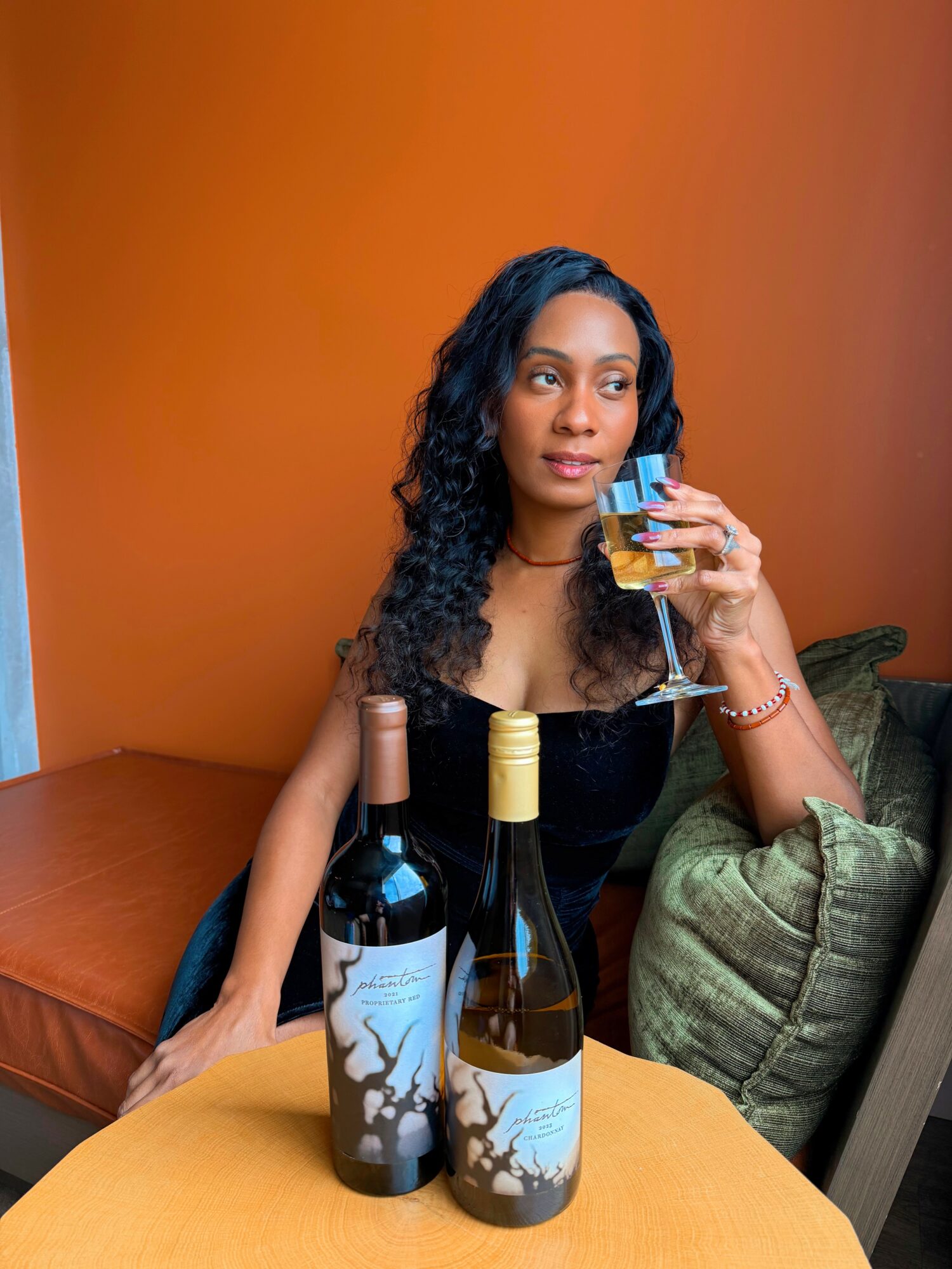 Woman with curly hair holding a glass of wine, sitting at a table with three wine bottles, in a cozy setting.