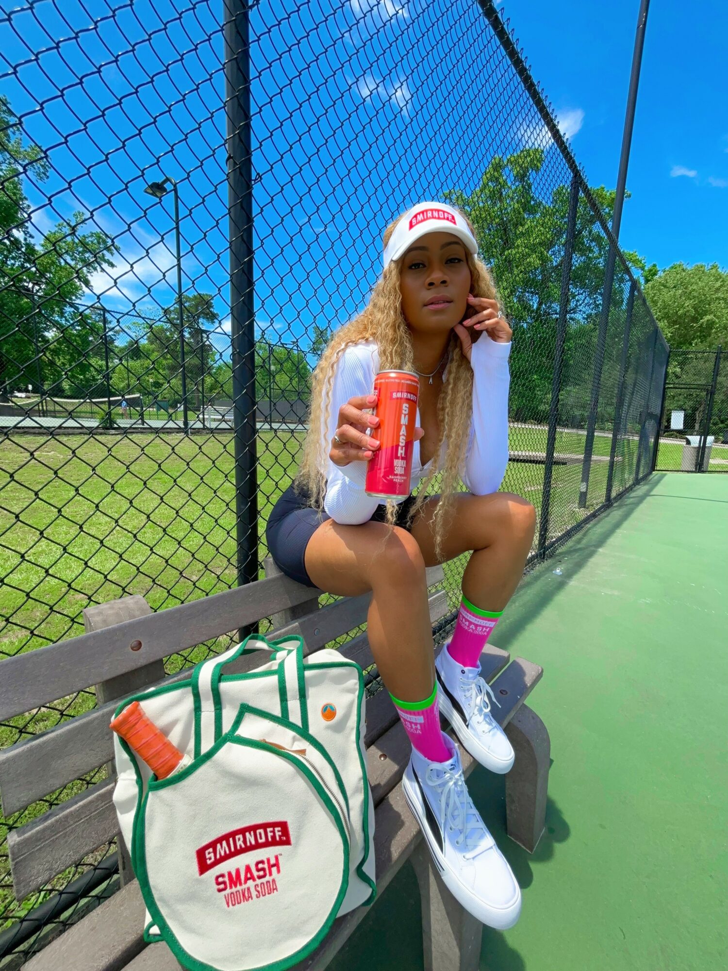 Young woman sitting on a bench at a tennis court, holding a drink, wearing a white cap, white top, black shorts, and pink socks.