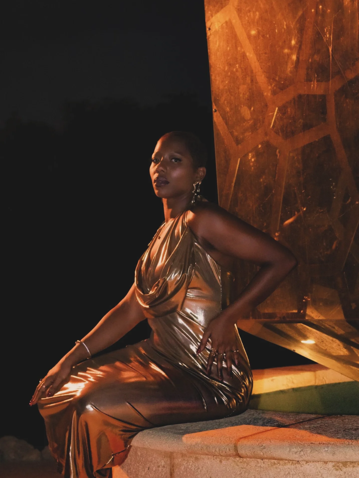Woman in a shiny gold dress sitting outdoors at night, illuminated by warm light, with a dark background and geometric structure.