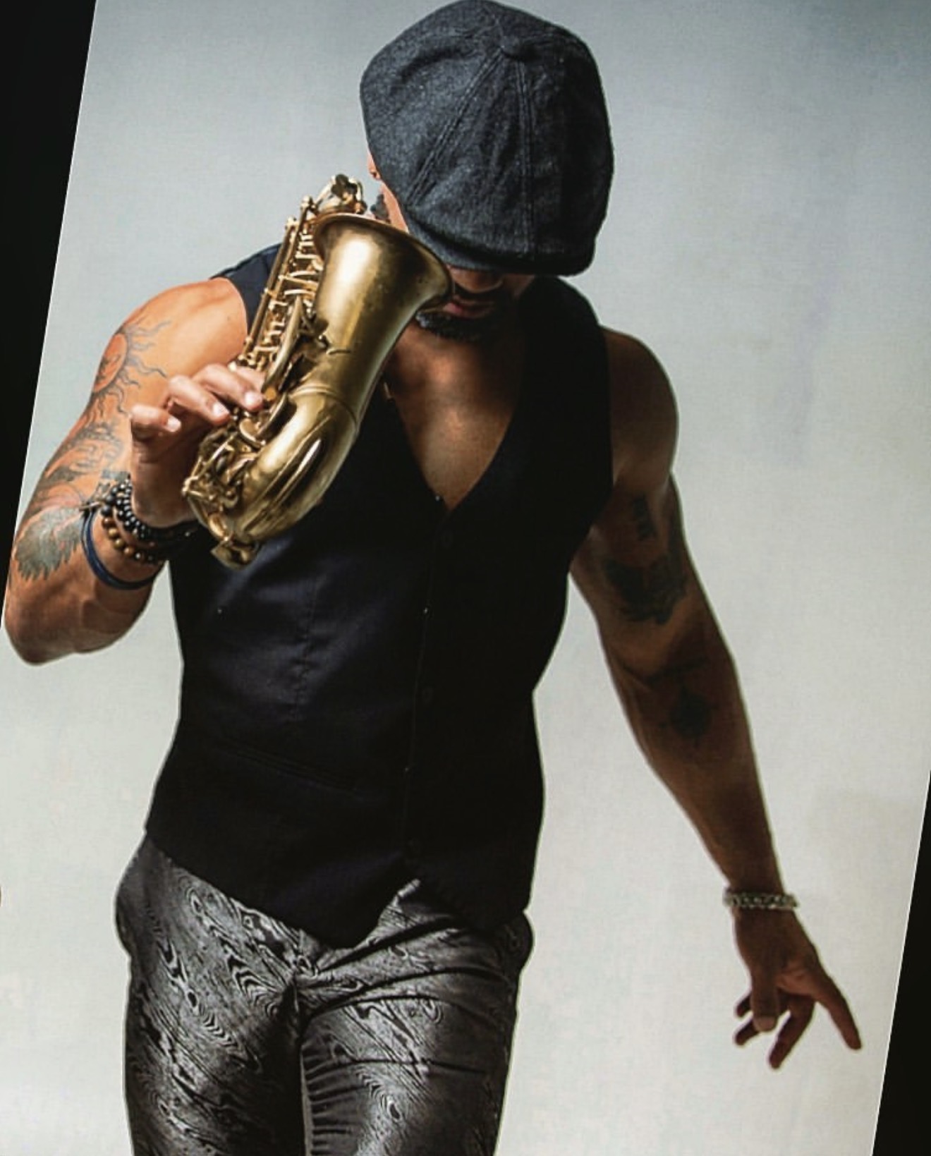 Person wearing a hat, sleeveless vest, holding a saxophone, with tattoos on arms, standing against a plain background.
