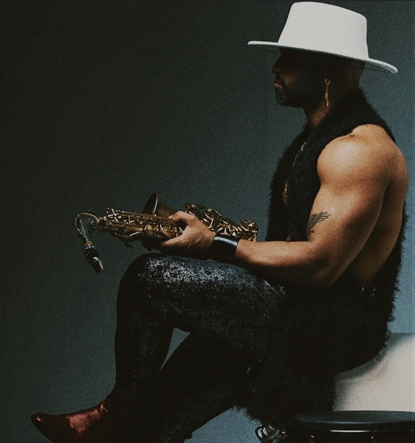 Person with a white hat, sitting shirtless, holding a saxophone, wearing dark pants, against a dark background.