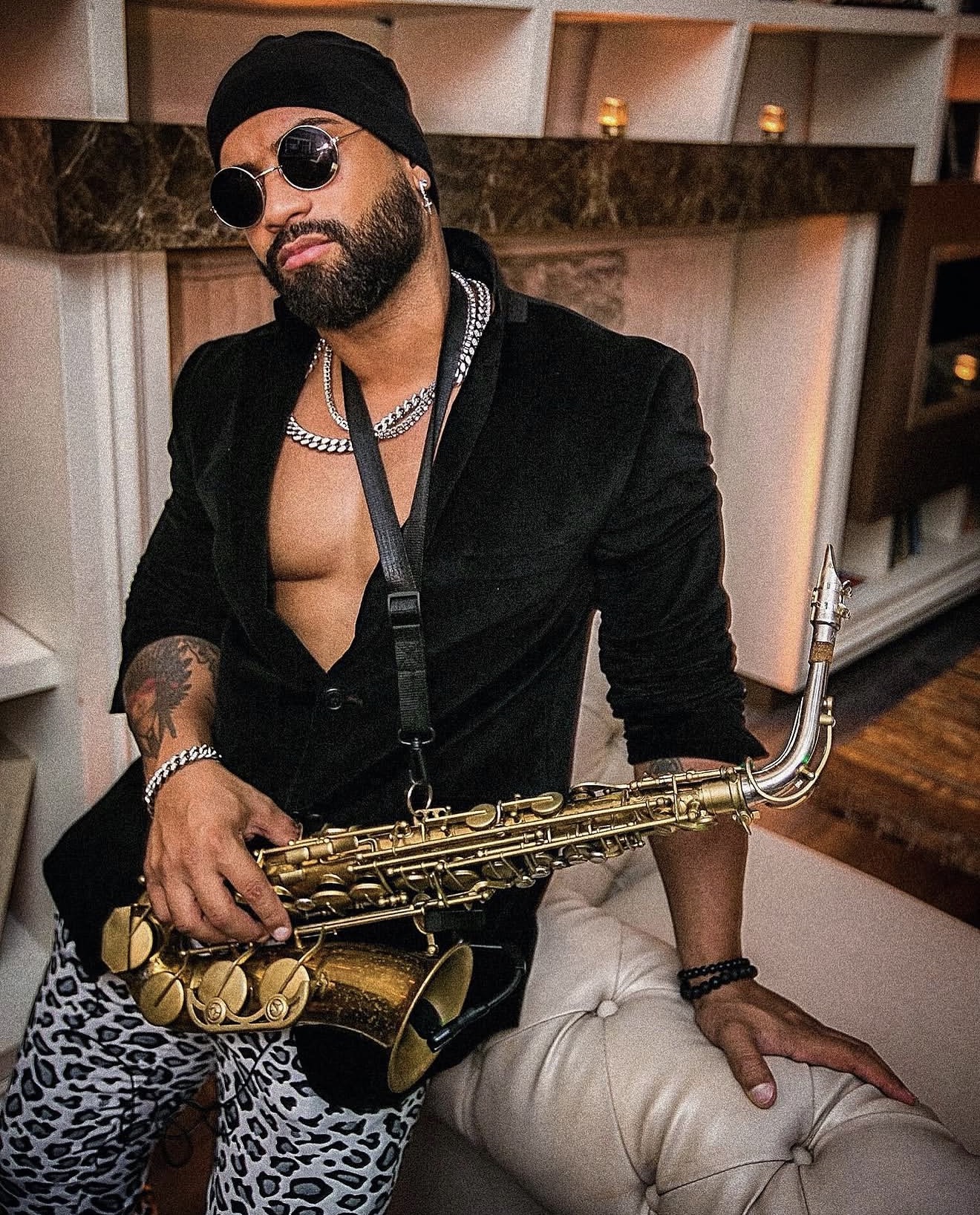 Man with sunglasses, beard, and tattoos holding a saxophone, wearing a black jacket and leopard print pants, sitting indoors.