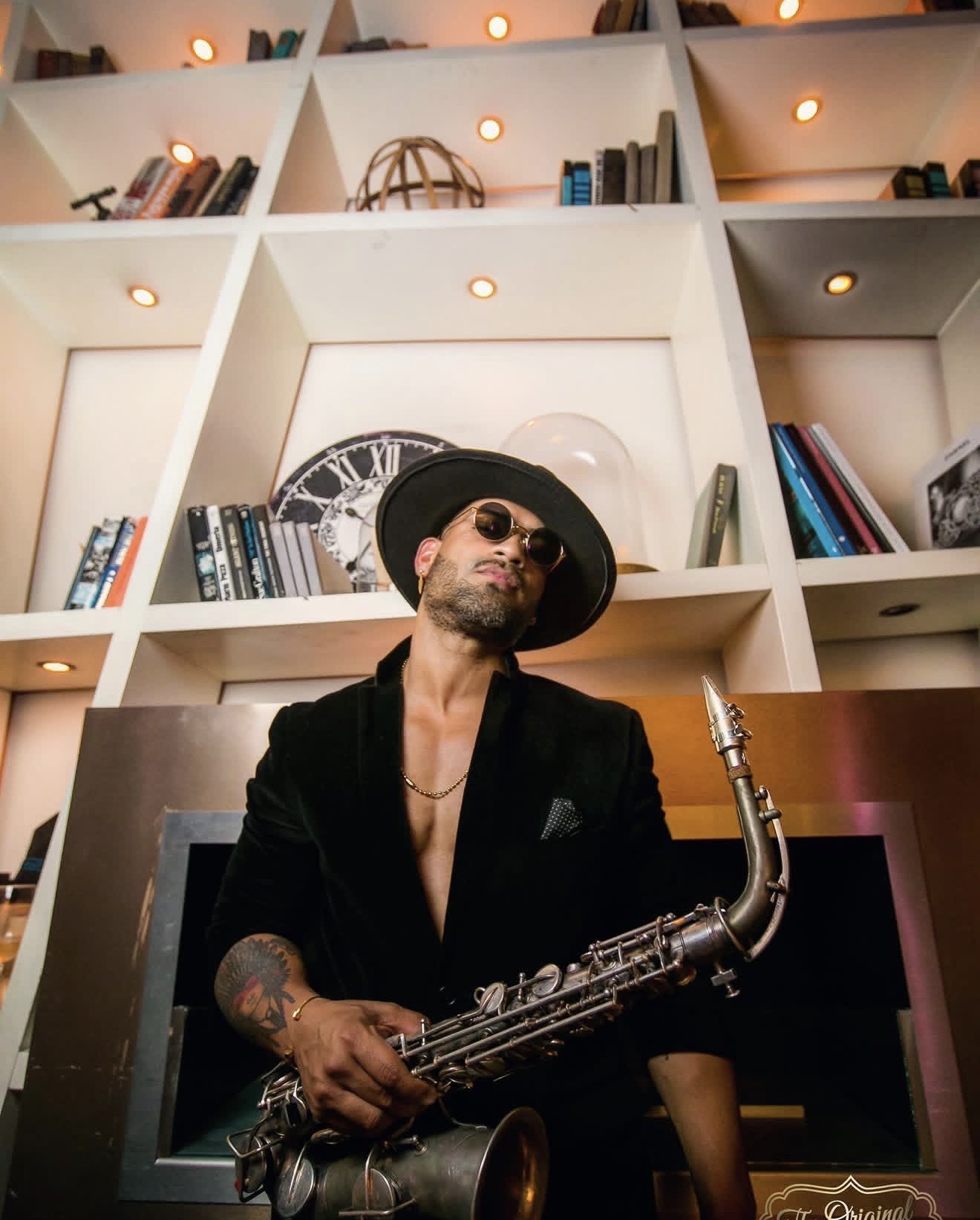 Man wearing sunglasses and a hat, holding a saxophone, sitting in front of a bookshelf with books and decorative items.