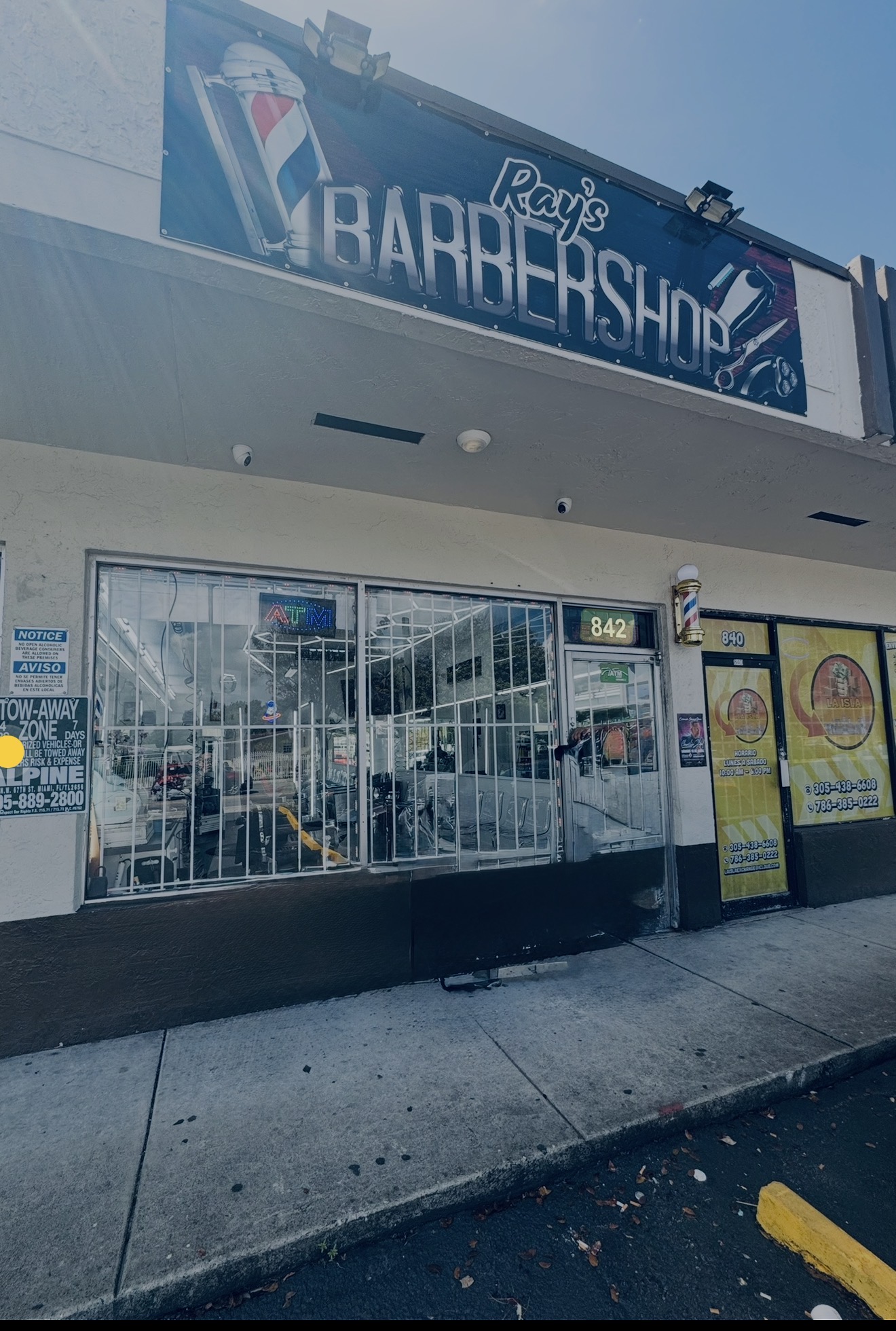 Barbershop storefront with large glass window, metal security bars, and signs outside, reflecting parked cars and buildings.