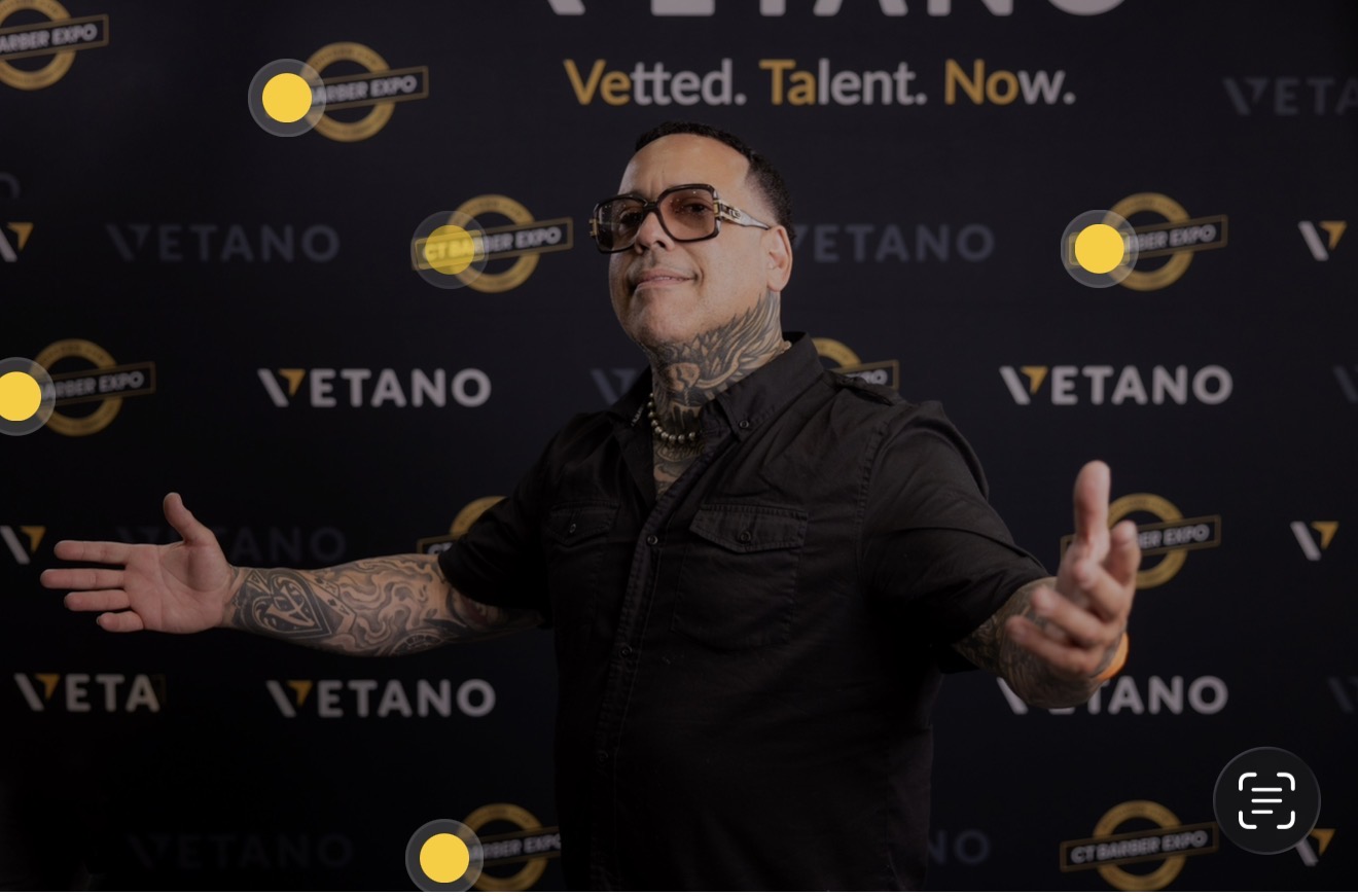 Person with tattoos and glasses standing with arms outstretched in front of a Vetano backdrop.