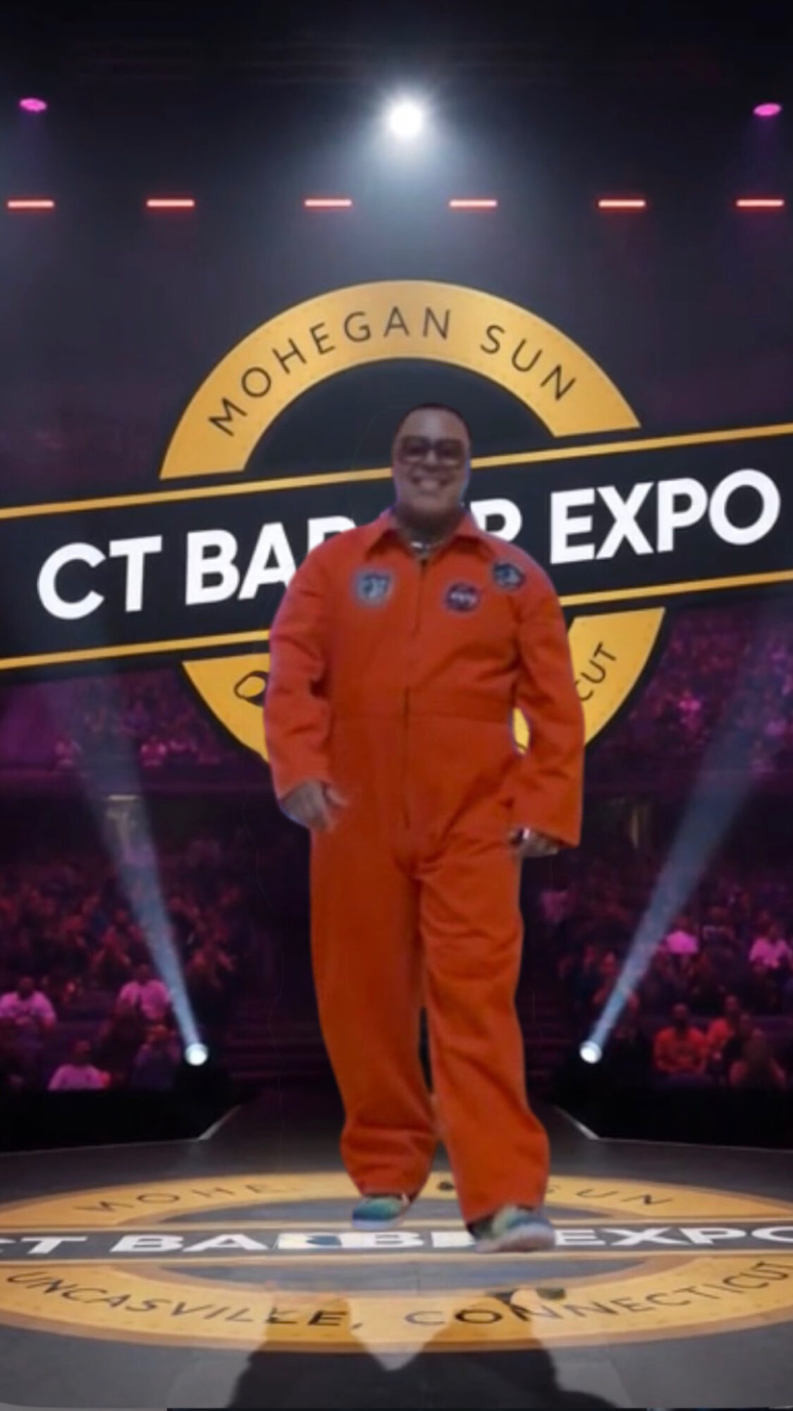 Person in orange jumpsuit standing on stage with a large circular logo behind them, stage lights shining down.