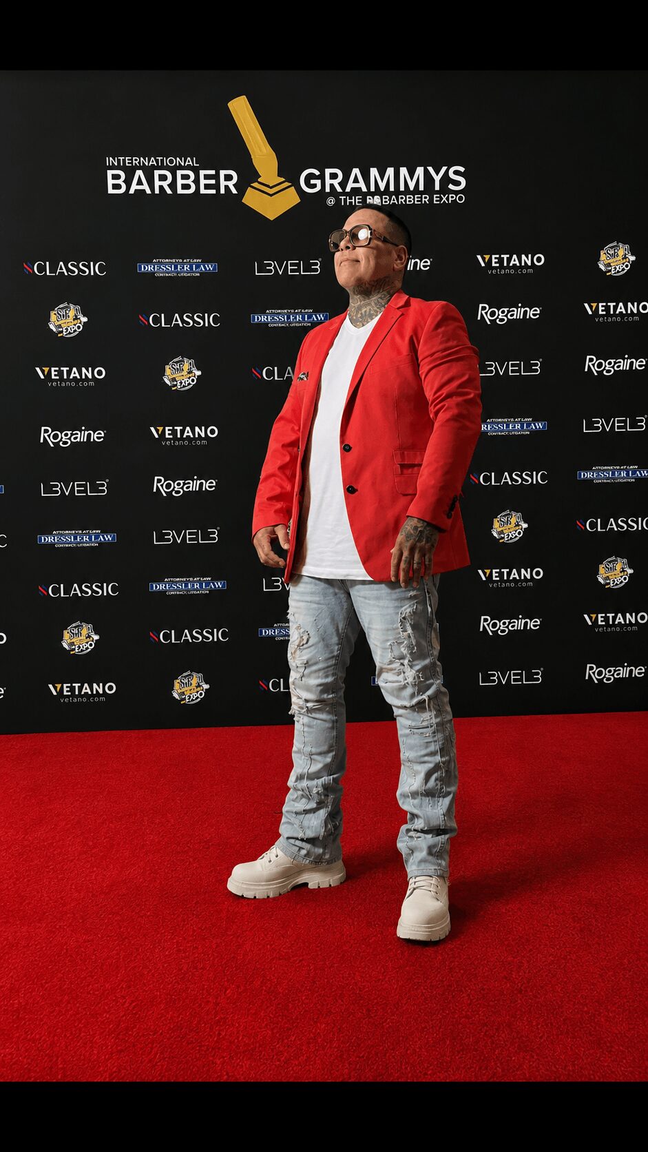 Man in red jacket and light jeans standing on red carpet at event backdrop, smiling, with hands in pockets.