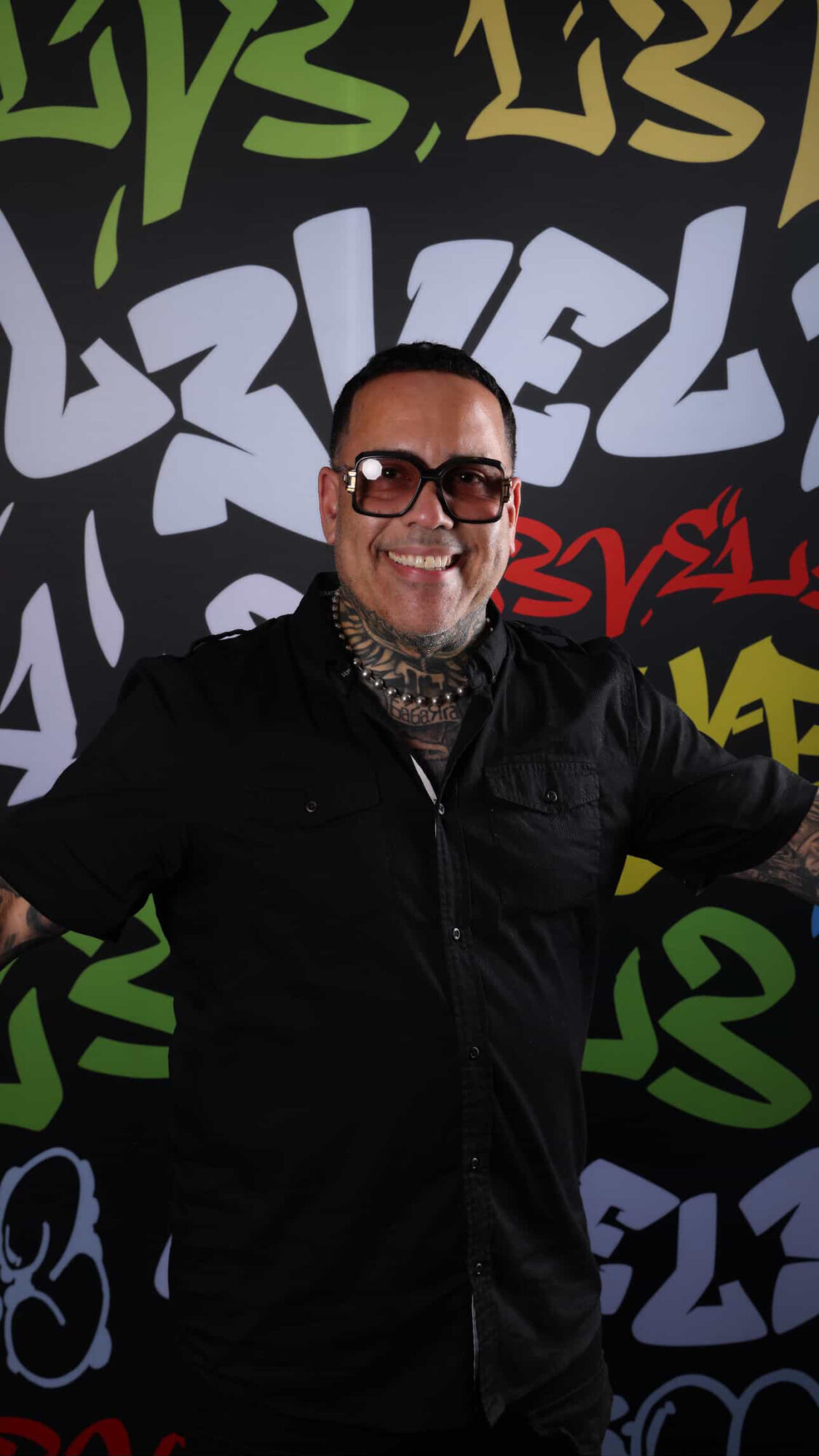 Person smiling wearing glasses and a black jacket, standing in front of colorful graffiti background.