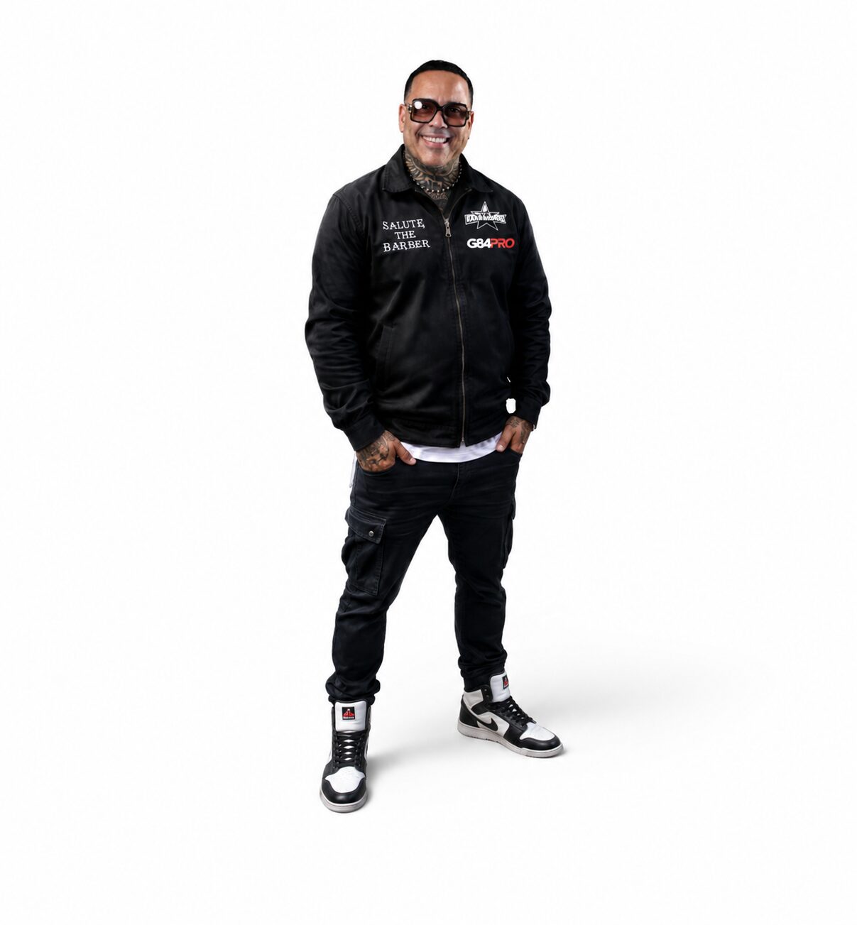 Man wearing sunglasses, black jacket, and sneakers, standing with hands in pockets, smiling, against a white background.