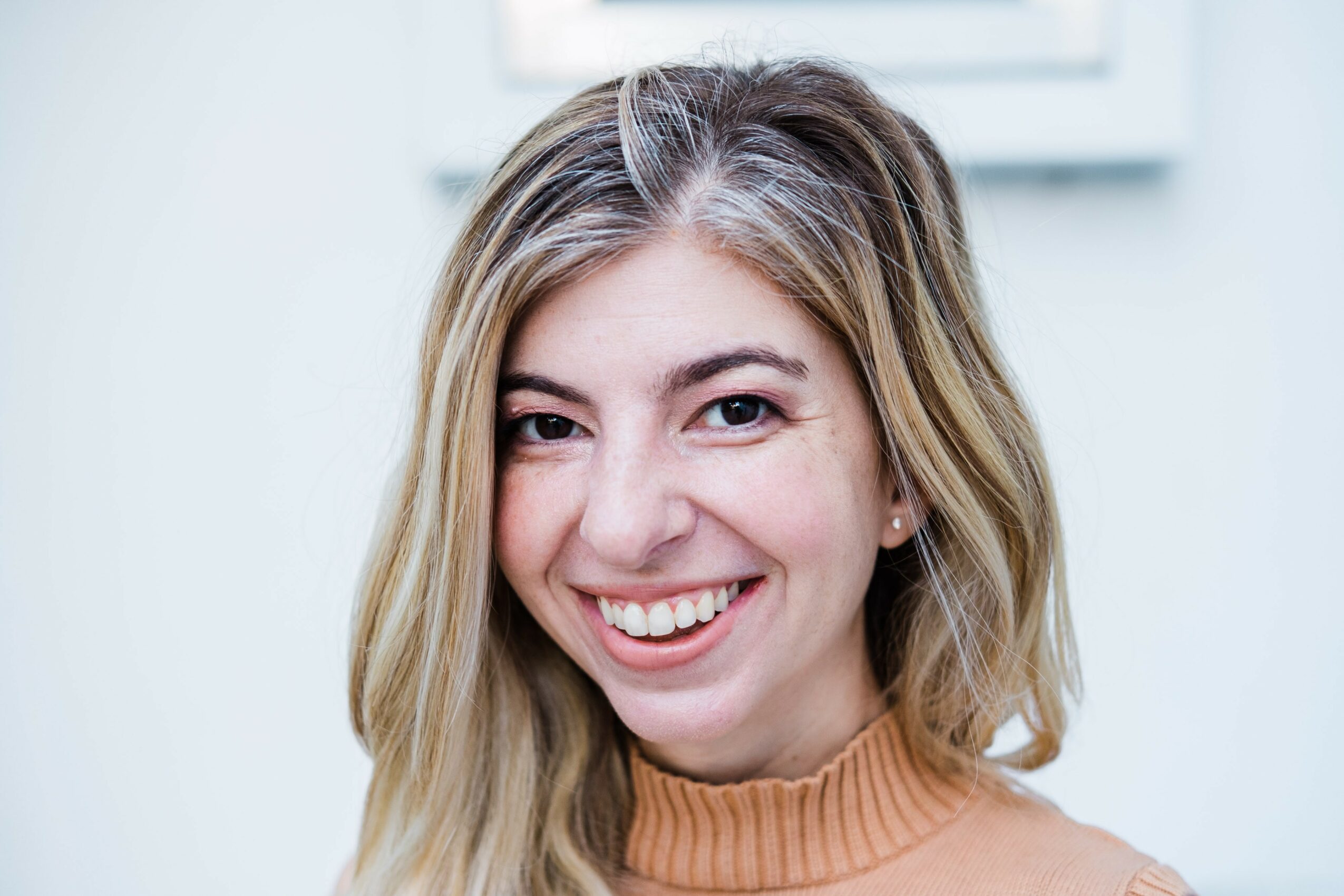 Young woman with shoulder-length blonde hair smiling, wearing a tan turtleneck, in a light-colored room.