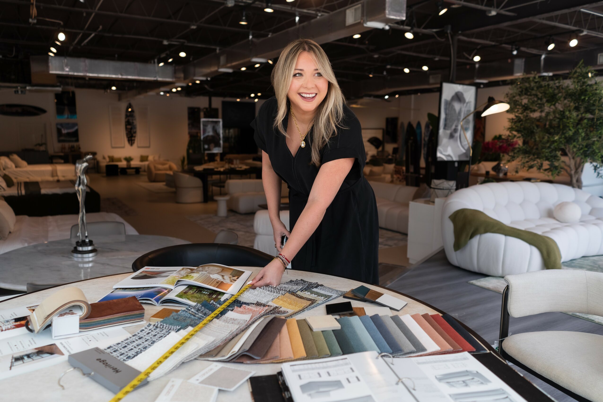 Woman with blonde hair smiling and leaning over a table with fabric samples in a furniture store.