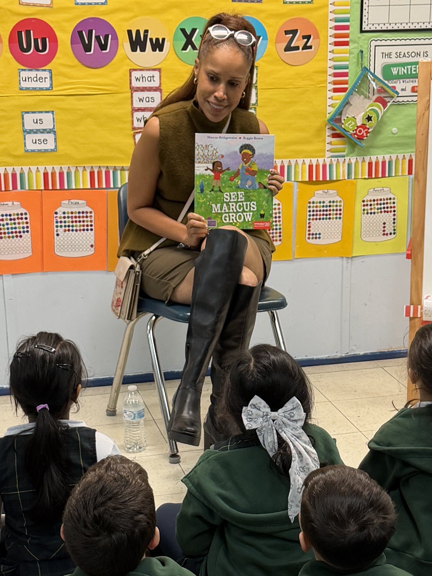 Woman reading a book to children in a classroom, colorful educational posters on the wall.