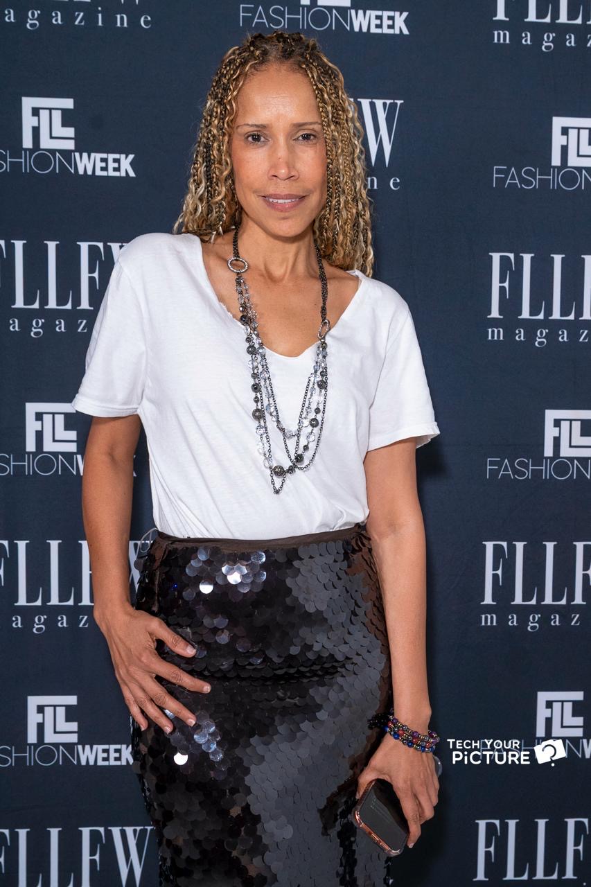 Woman with curly hair in white top and black sequined skirt at fashion event, holding a phone, standing in front of a blue backdrop.