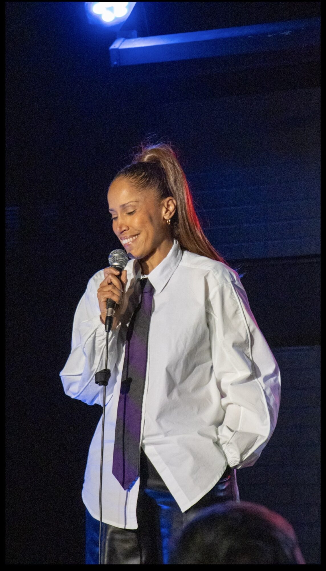 Young woman with a ponytail smiling, holding a microphone on stage, wearing a white shirt and purple tie.
