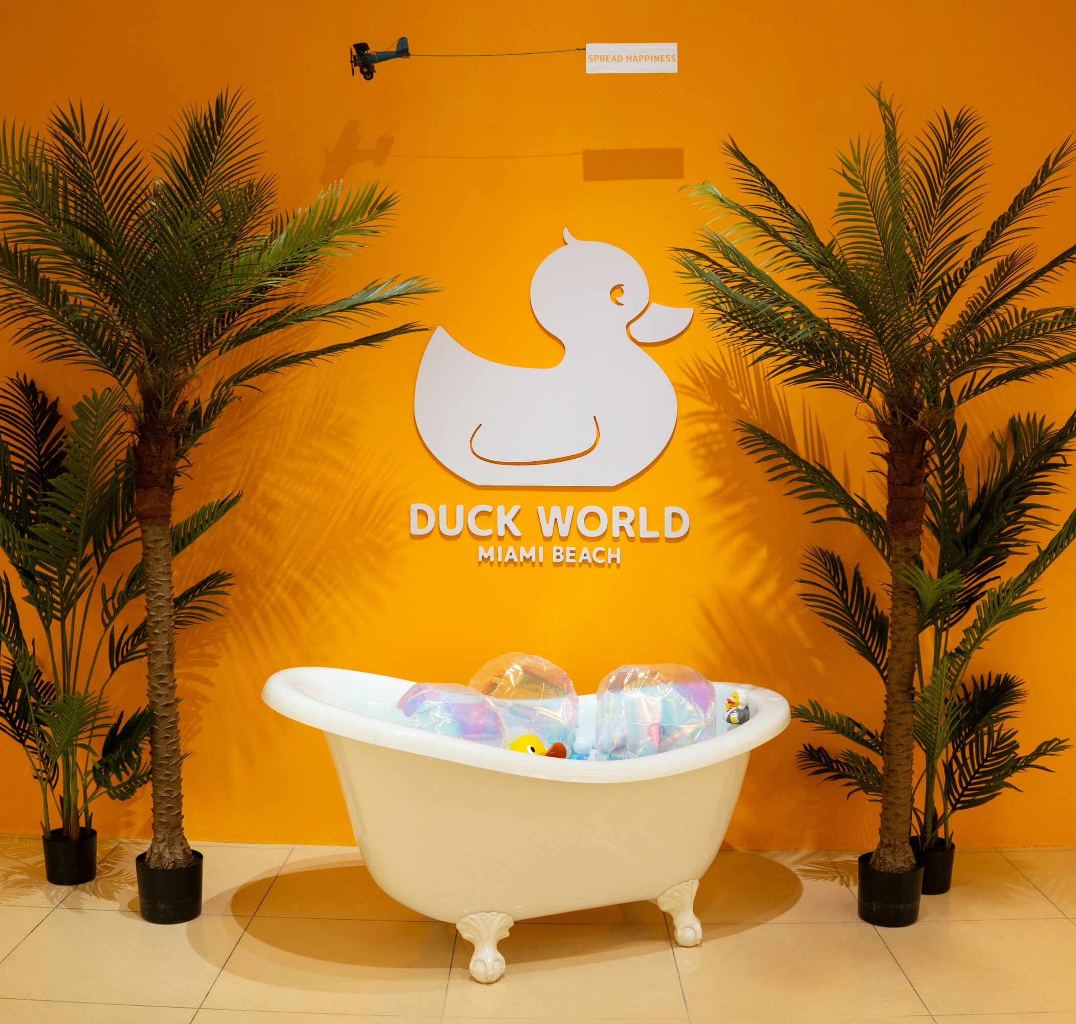 White bathtub filled with colorful plastic balls, flanked by two potted palm trees against an orange wall with a duck logo.