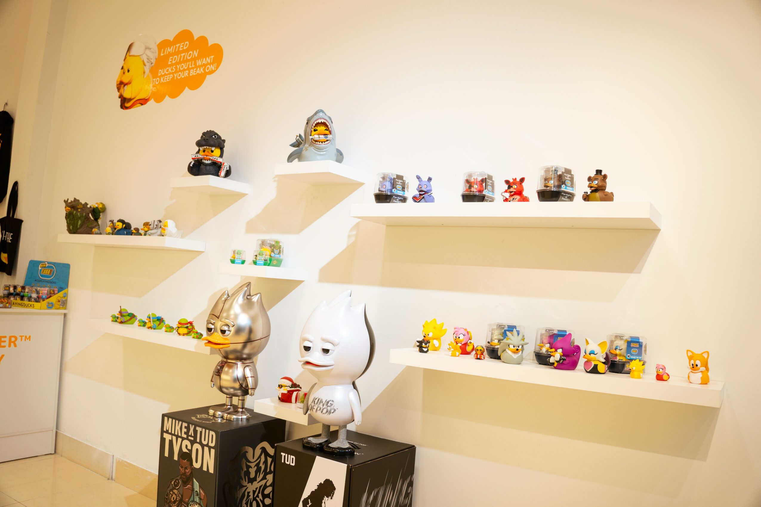 Display of various small toys and figures on white shelves against a white wall, with two larger figures in front, one with a metallic head.