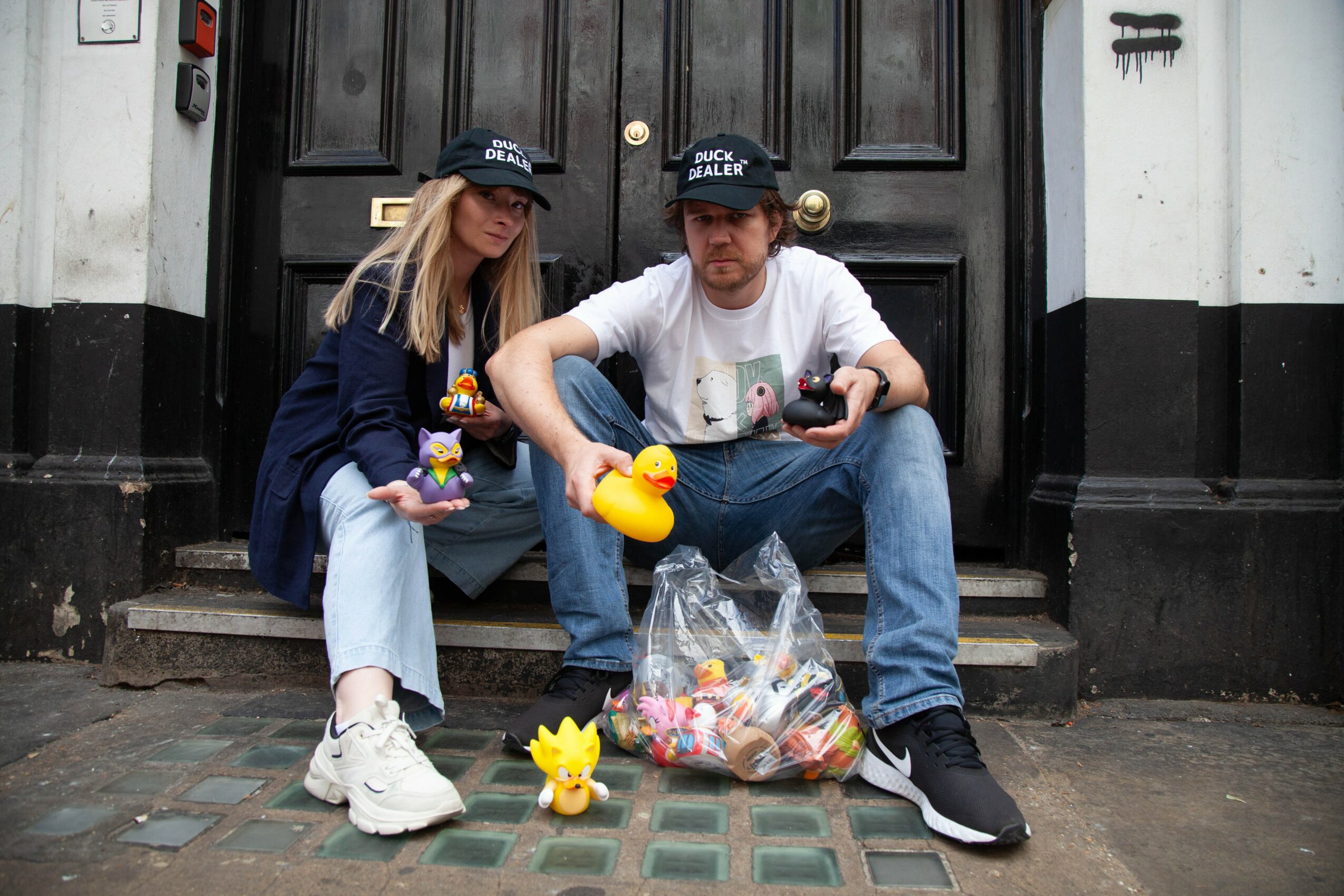 Two young people sit on steps in front of a black door, holding rubber ducks and a bag of toys, with a yellow duck on the ground.