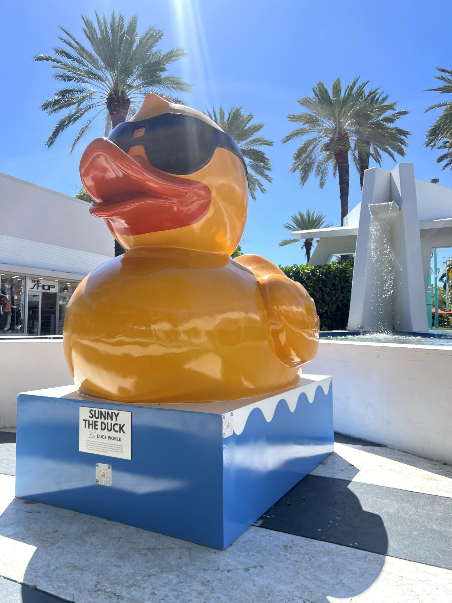 Large rubber duck sculpture wearing sunglasses, outdoors with palm trees and blue sky, on a blue platform.