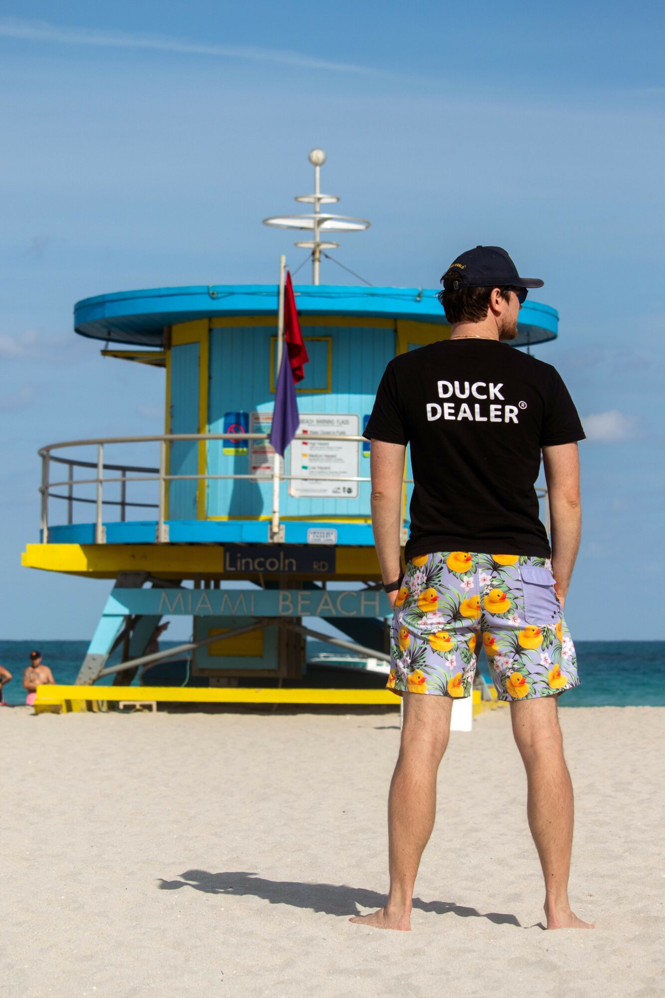 Person wearing a black cap and T-shirt with 'DUCK DEALER' standing on sandy beach near a lifeguard tower, ocean in background.