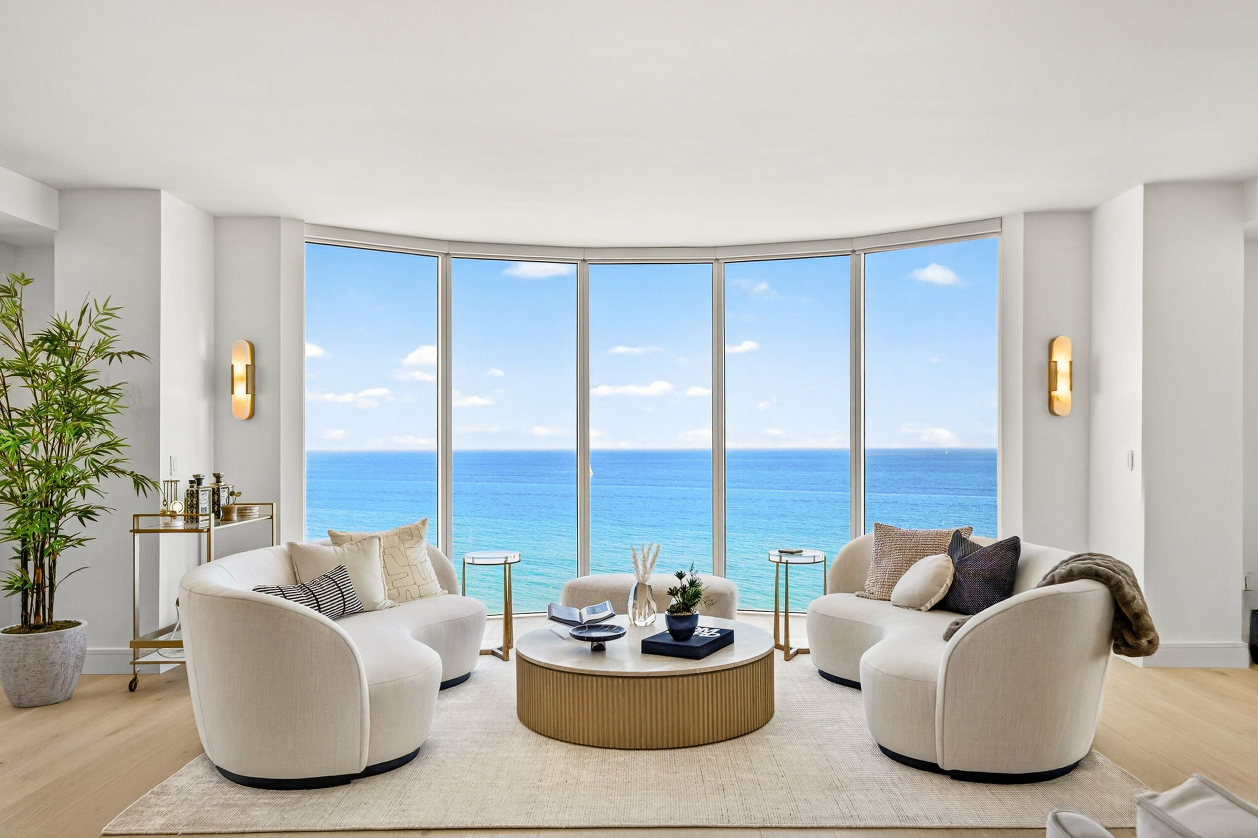 Living room with large windows showing ocean view, two white sofas, a round coffee table, and indoor plants.