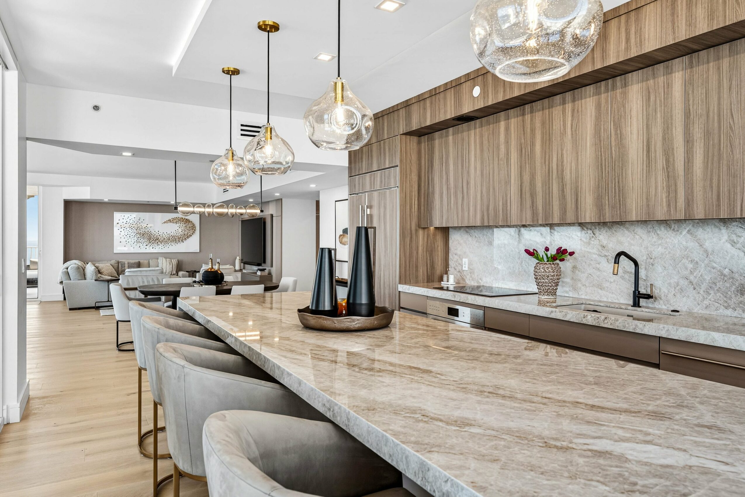 Modern kitchen with a marble countertop, wooden cabinets, pendant lights, and a sink with a potted plant, open to living area.
